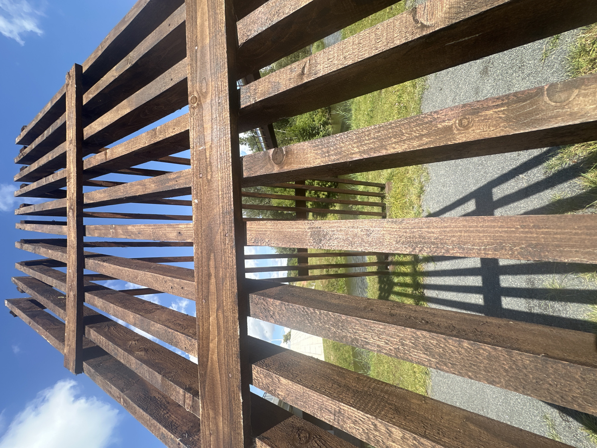 An outdoor, low-angle, wide shot shows a wooden structure made of dark brown planks under a bright blue sky with scattered white clouds. The structure appears to be a gate or part of a fence, constructed with vertical and horizontal wooden beams. Sunlight casts a shadow of the wooden slats onto the ground, which is a mix of gray gravel and green grass. In the background, a body of water, possibly a river or canal, is visible, lined with green trees. A white concrete structure and a bridge are also faintly visible in the distance. The image is taken at the National Library of Belarus in Minsk, Belarus.