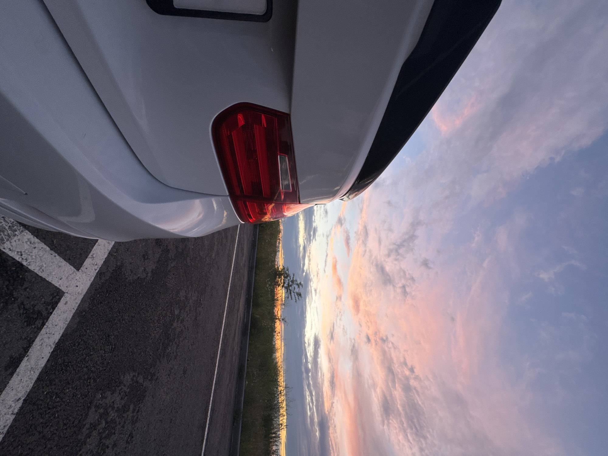 The image captures a close-up view of the rear of a white car, specifically the taillight and part of the trunk. The car is parked on asphalt, with white parking lines visible in the foreground. To the right of the car, the scene opens up to a broad landscape under a dramatic sky at sunset. The sky is filled with clouds tinged with soft pinks, oranges, and purples, indicative of the golden hour. A thin strip of vegetation, including some sparse trees and grass, separates the road from the sky. The overall mood is serene and picturesque, highlighting the natural beauty of the sunset against the man-made object. There is no visible text in the image. The location context suggests this scene is in Mikhaylovsk, Russia.