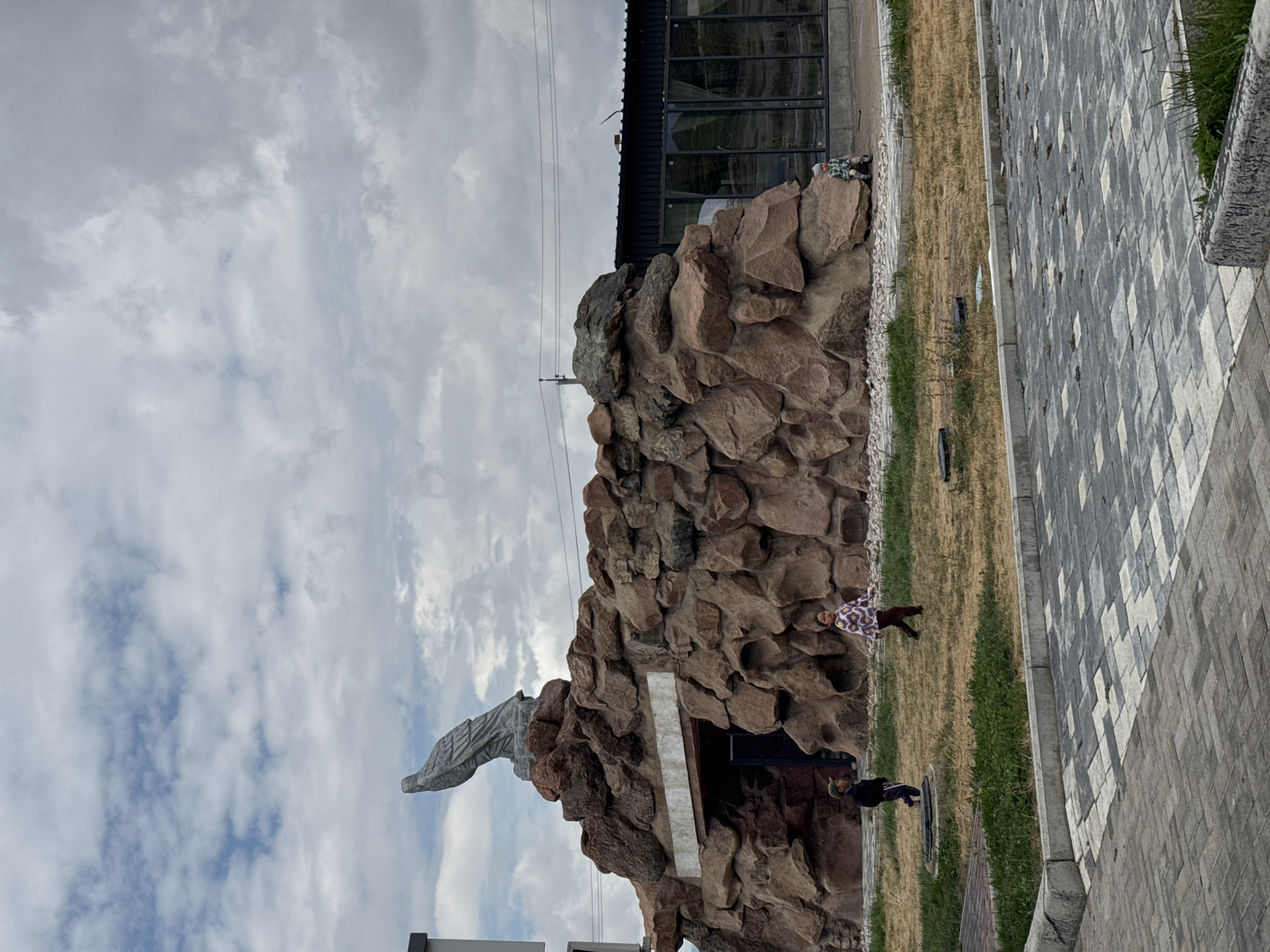 In Balykchy, Kyrgyzstan, on a cloudy day, a stone statue of a bird of prey perches atop a man-made rock formation that resembles a cave entrance. To the left of the formation, a young boy wearing a cap and dark clothing walks on dry grass. To the right of the formation, a young girl in a patterned top and dark pants walks across the grass. Further to the right, a child in camouflage clothing sits on a rock. Behind the rock formation, a modern building with large glass windows is visible, suggesting a commercial or public establishment. The foreground features a paved area with a patterned design of grey and white tiles. The overall scene depicts an outdoor public space with a decorative element and children present.