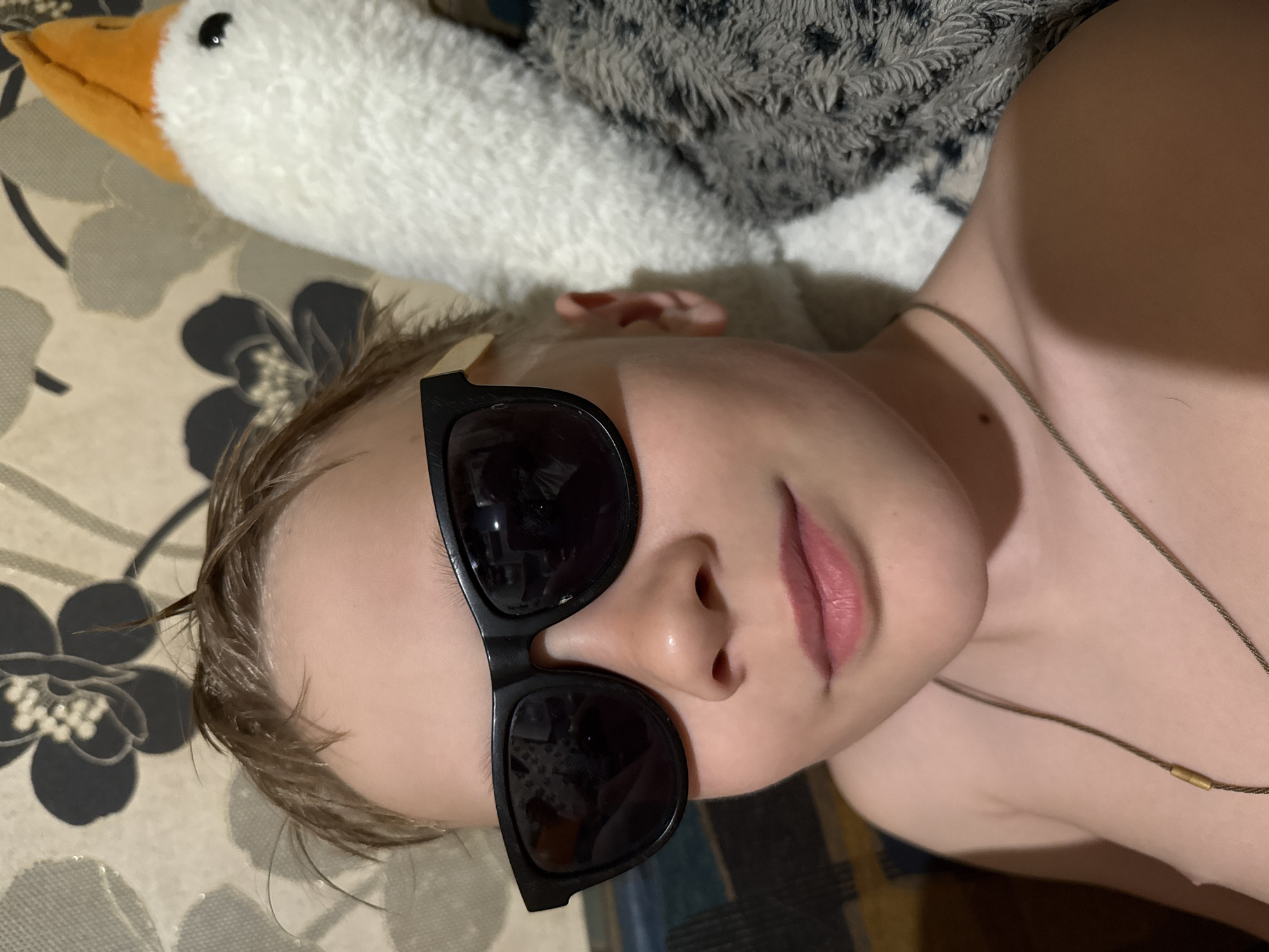 A fair-skinned child wearing dark sunglasses lies on a patterned surface. A plush toy resembling a goose is visible in the background. The child wears a thin necklace. The location in the city of Dmitrov, Russia, is not ascertainable from the given visual information.