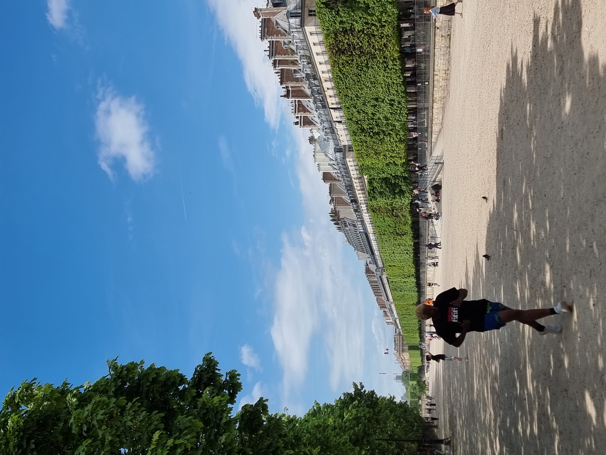 The image shows a sunny day in the Tuileries Garden in Paris, France. The camera is rotated 90 degrees counter-clockwise. A person with blonde hair, wearing a black t-shirt and blue shorts is walking down a gravel path. Buildings line the path behind a wall of trees and greenery. Other people are seated along the path. There are trees at the bottom of the frame and a bright blue sky with white clouds above. Tree shadows are visible on the gravel path.