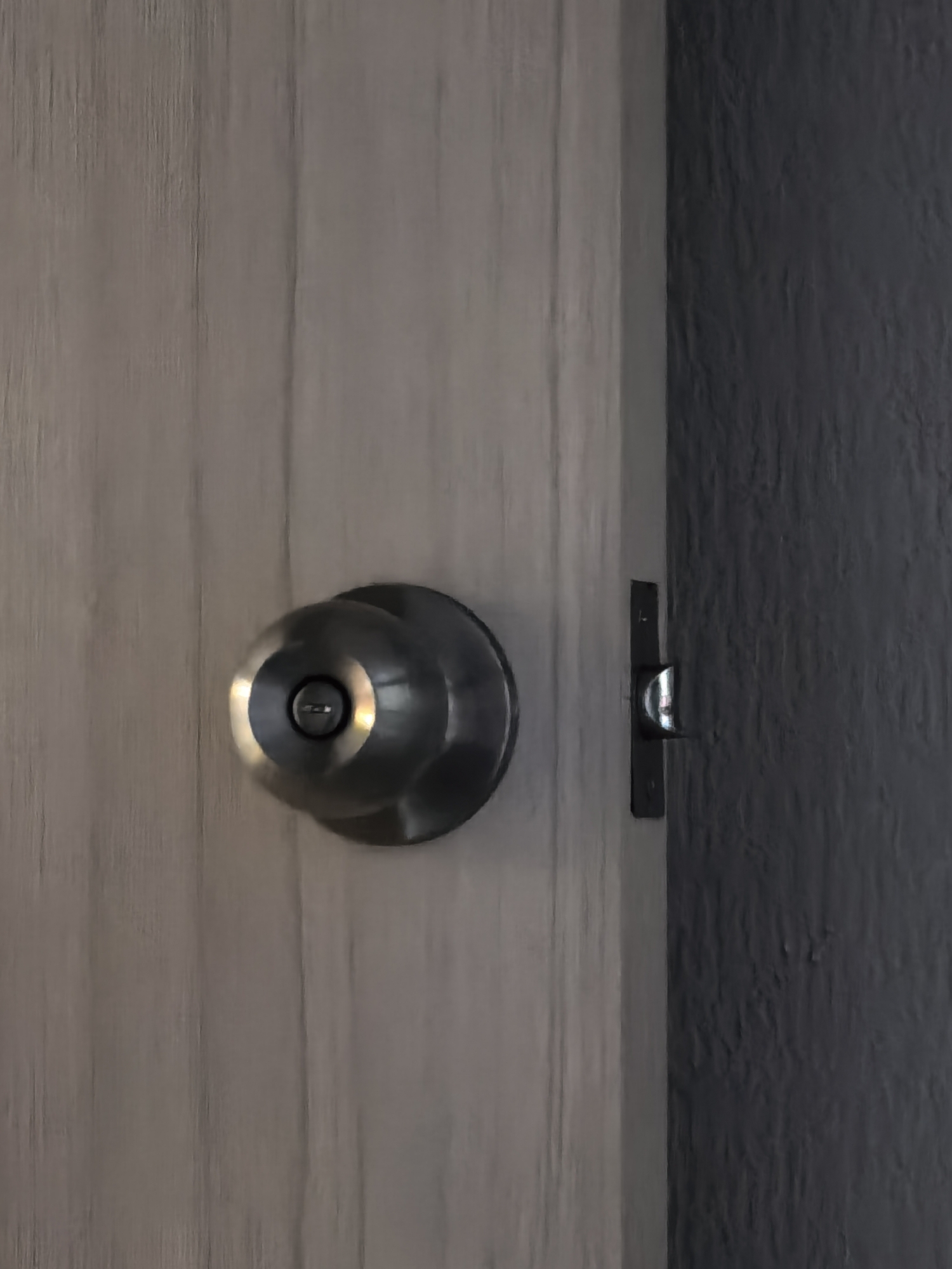 The image shows a close-up of a door handle and lock mechanism. The door handle is a round, silver, metallic knob with a keyhole in the center. It is mounted on a light-colored wooden door with a visible wood grain pattern. To the right of the door handle, a metal latch is visible, extending from a dark-colored wall into the edge of the wooden door. The dark wall has a textured surface. The lighting suggests an indoor setting, with reflections visible on the metallic door handle. The image focuses on the details of the door hardware and the transition between the two different wall surfaces. There is no visible text, indication of time of day, weather, or any human presence. The overall scene is static and focuses on architectural elements.