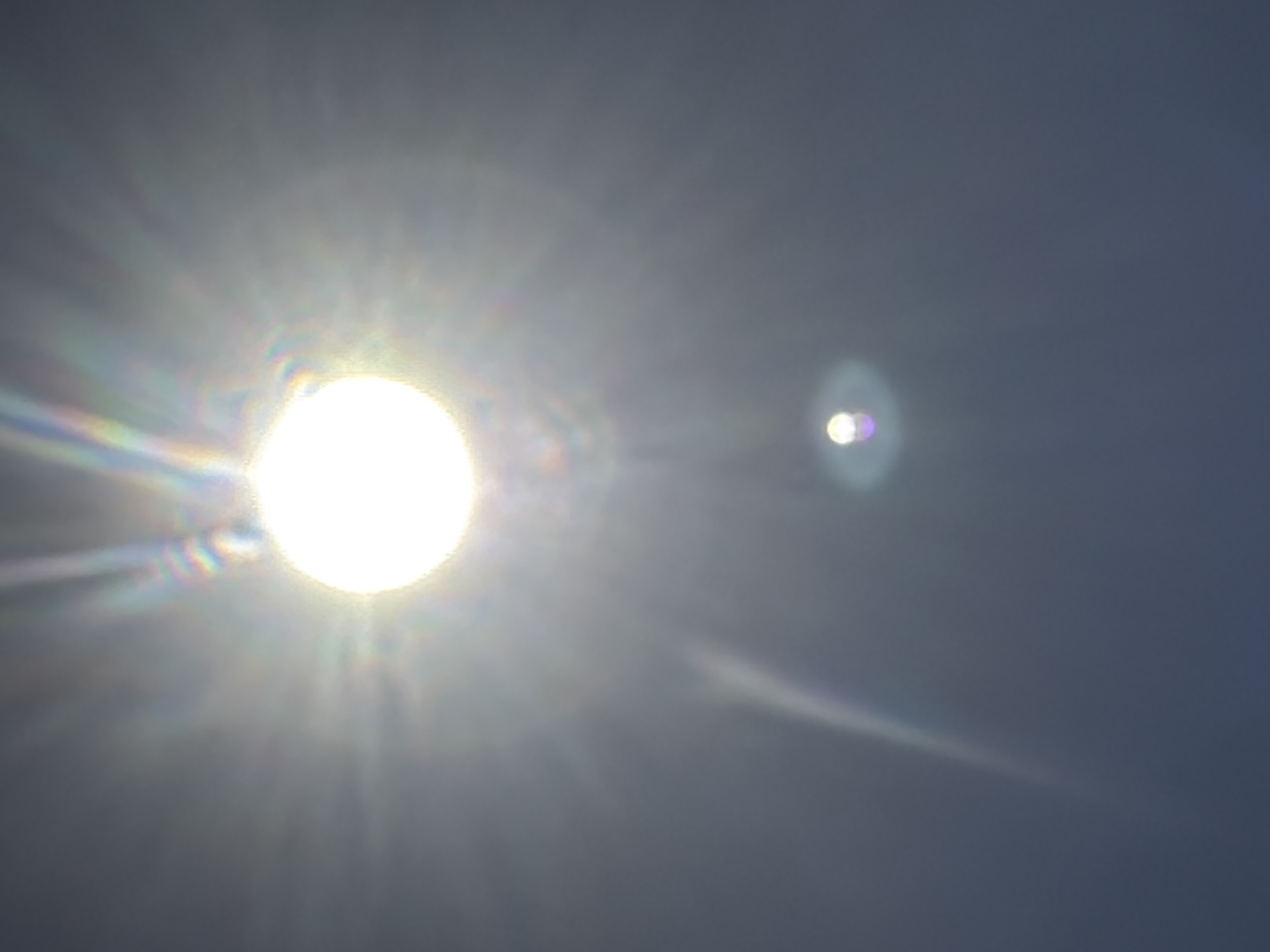 The image shows a bright, circular light source, likely the sun, in the center of the frame. The light is intensely bright, with visible rays emanating outwards and creating a lens flare effect. There are also rainbow-like colors visible within the flare, suggesting the presence of light refraction. Below the primary light source, there is a smaller, fainter circular light, which appears to be another lens flare or a reflection. The background is a dark, out-of-focus sky. The scene appears to be taken outdoors, possibly during the day, given the brightness of the light. The image does not contain any identifiable landmarks or objects that would indicate a specific location within Sharm El Sheikh, Egypt.