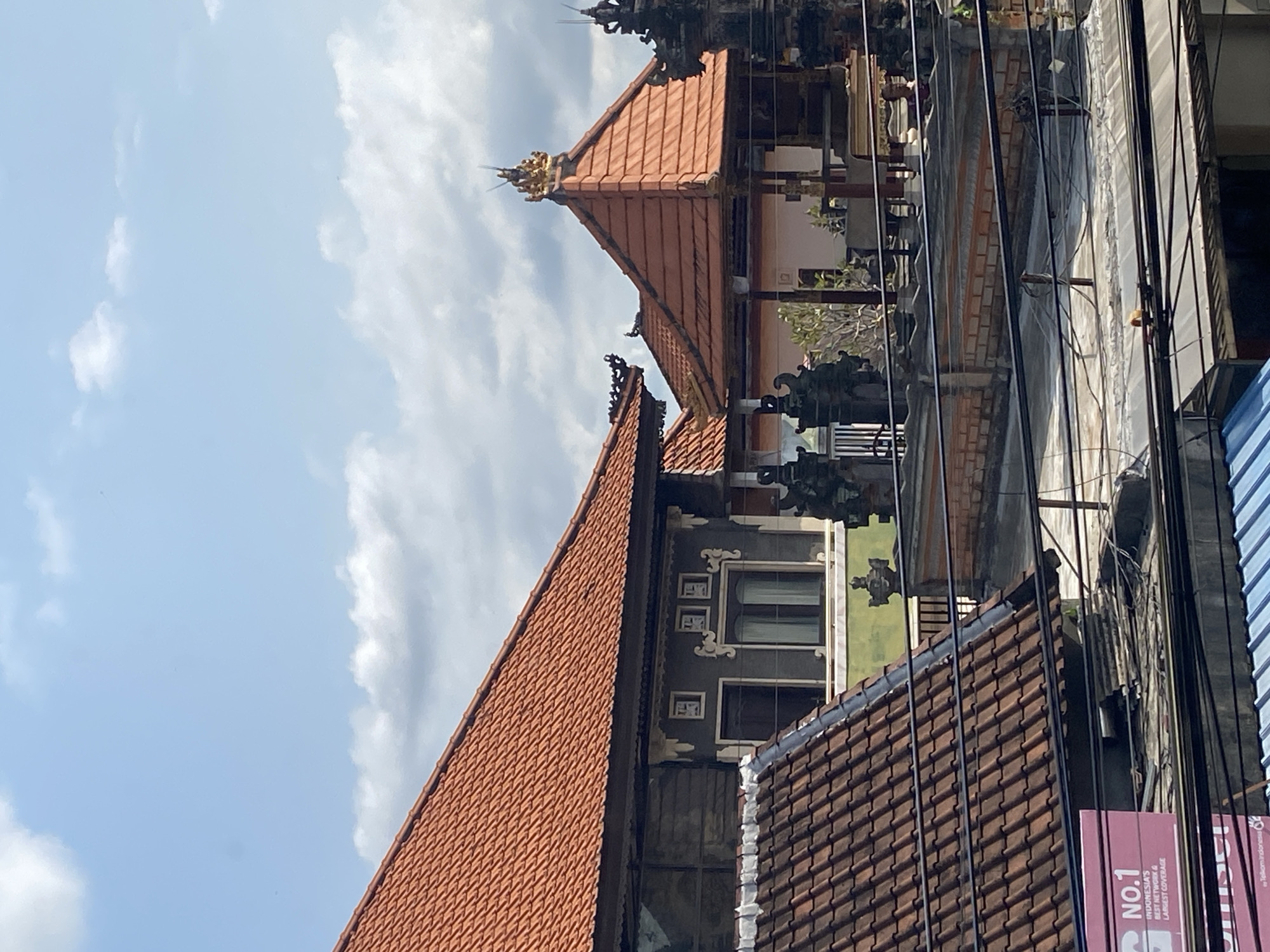 In North Kuta, Indonesia, the image presents a low-angle view of traditional Balinese architecture set against a sky dotted with light, fluffy clouds. The primary subjects are tiered roofs with distinct red clay tiles, indicative of local construction. Architectural details such as intricate carvings and embellishments are visible on the rooflines. Power lines cut through the scene, a common feature of urban environments. A building with a gray exterior and rectangular windows stands adjacent to the traditional structure. An advertisement featuring the text "NO.1 INDONESIA'S NETWORK & LARGEST COVERAGE" is visible on the lower right of the image.