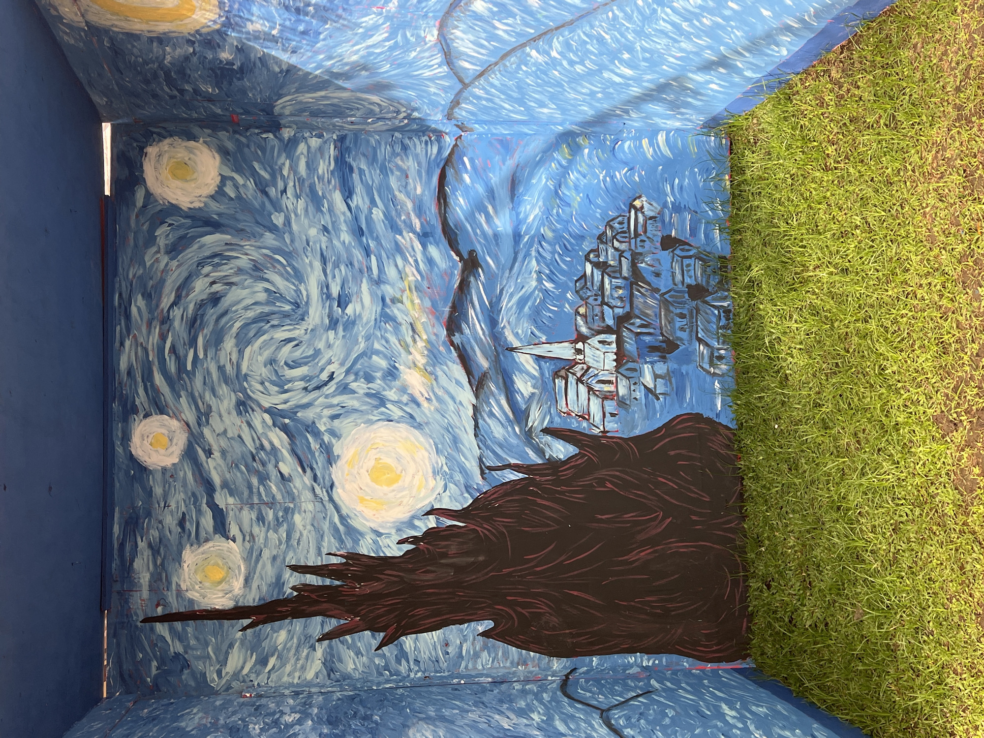 The image shows a three-sided mural painted to resemble Vincent van Gogh's "The Starry Night." The mural is on the interior walls of what appears to be a small, open-air structure. The walls are painted with swirling blue and white brushstrokes representing the night sky, with yellow circles for stars and a crescent moon. A large, dark, spiky tree is painted on the left wall, and a village with a church spire is depicted in the center. The floor of the structure is covered with green grass. The structure is located in the city of North Kuta, Indonesia.
