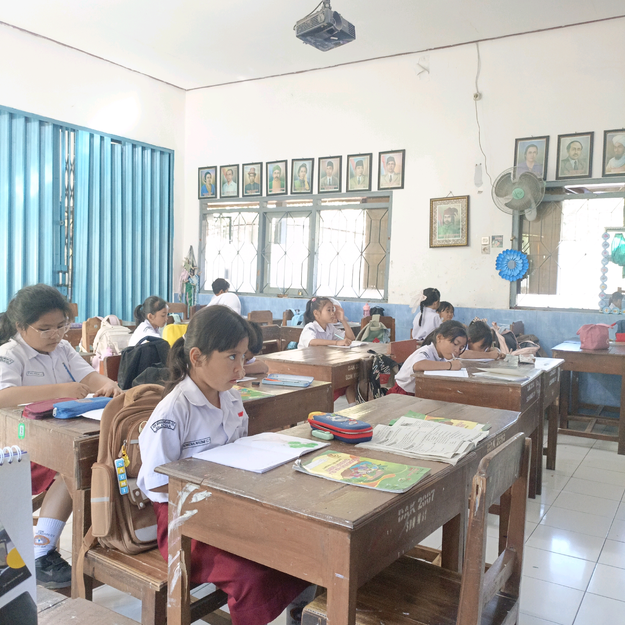 The image depicts a classroom scene at a school in Salatiga, Indonesia. Several young girls, dressed in school uniforms, are seated at wooden desks, engaged in learning activities. The classroom environment is simple, with basic wooden furniture and a light-colored tiled floor. The walls are adorned with framed portraits of distinguished individuals, suggesting a historical or patriotic theme. A projector is mounted on the ceiling, and a ceiling fan is visible. The room is brightly lit, with natural light entering from windows. One student in the foreground is focused on writing in a notebook, while others are shown reading or looking towards the front of the classroom. The overall impression is one of an active and traditional educational setting in Salatiga.