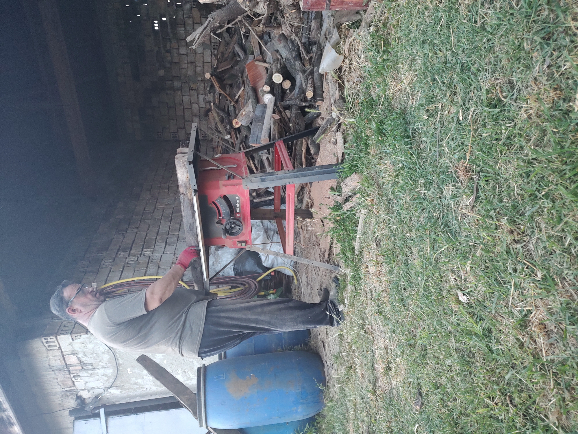 This media file features a man standing in an outdoor, rustic setting. He is wearing glasses, a grey t-shirt, dark pants, and red gloves. The man is positioned next to a red table saw with a circular blade. He appears to be engaged in a task involving the saw and some wooden materials. The background is a jumble of cut wood and what looks like a partially constructed brick structure. To the left, there's a dark, shadowy area, possibly an open shed or building. To the right and in the foreground, there's a patch of grass, some of which is dry and yellowish, suggesting it might be a rural or backyard area. Two large blue barrels are visible in the lower right portion of the image, one of which is partially obscured by the man's legs. The overall impression is one of a DIY project or outdoor work. The lighting suggests it is daytime, and the weather appears to be fair. The scene is set in Kochmar, Bulgaria. Visible text on the table saw includes "Einhell".