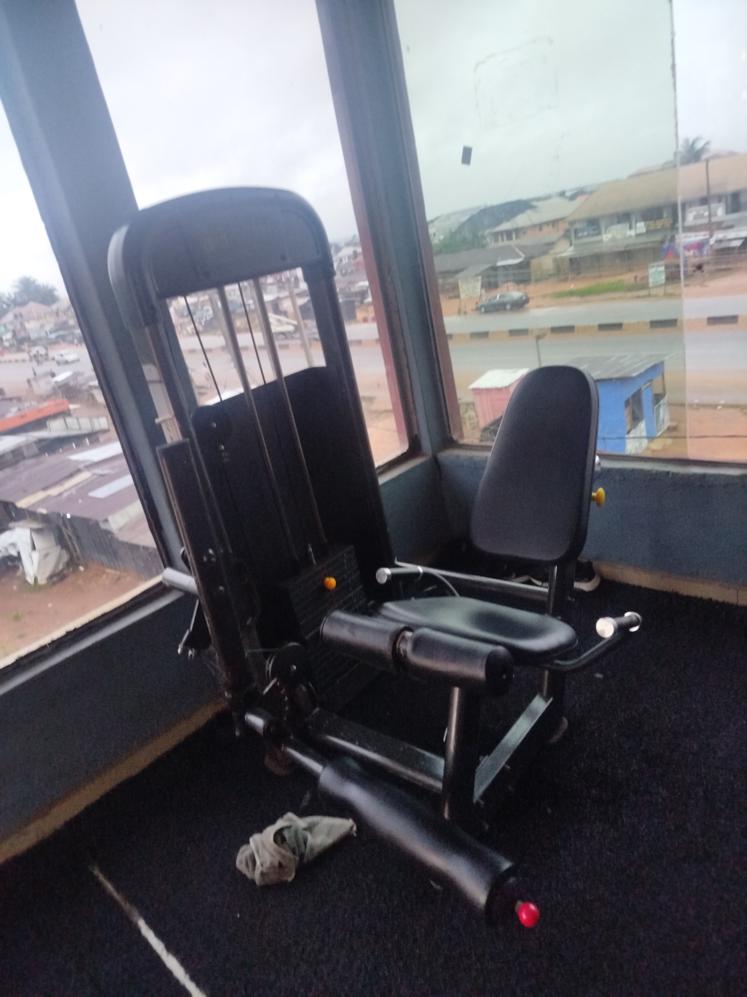 In Uselu, Nigeria, a black weight machine with a padded seat and backrest is positioned in a gym with large windows overlooking a street. A small, light-colored rag lies on the black carpeted floor in front of the machine. Outside, buildings and vehicles are visible, suggesting an urban environment. The lighting is dim, indicating an overcast or indoor setting.