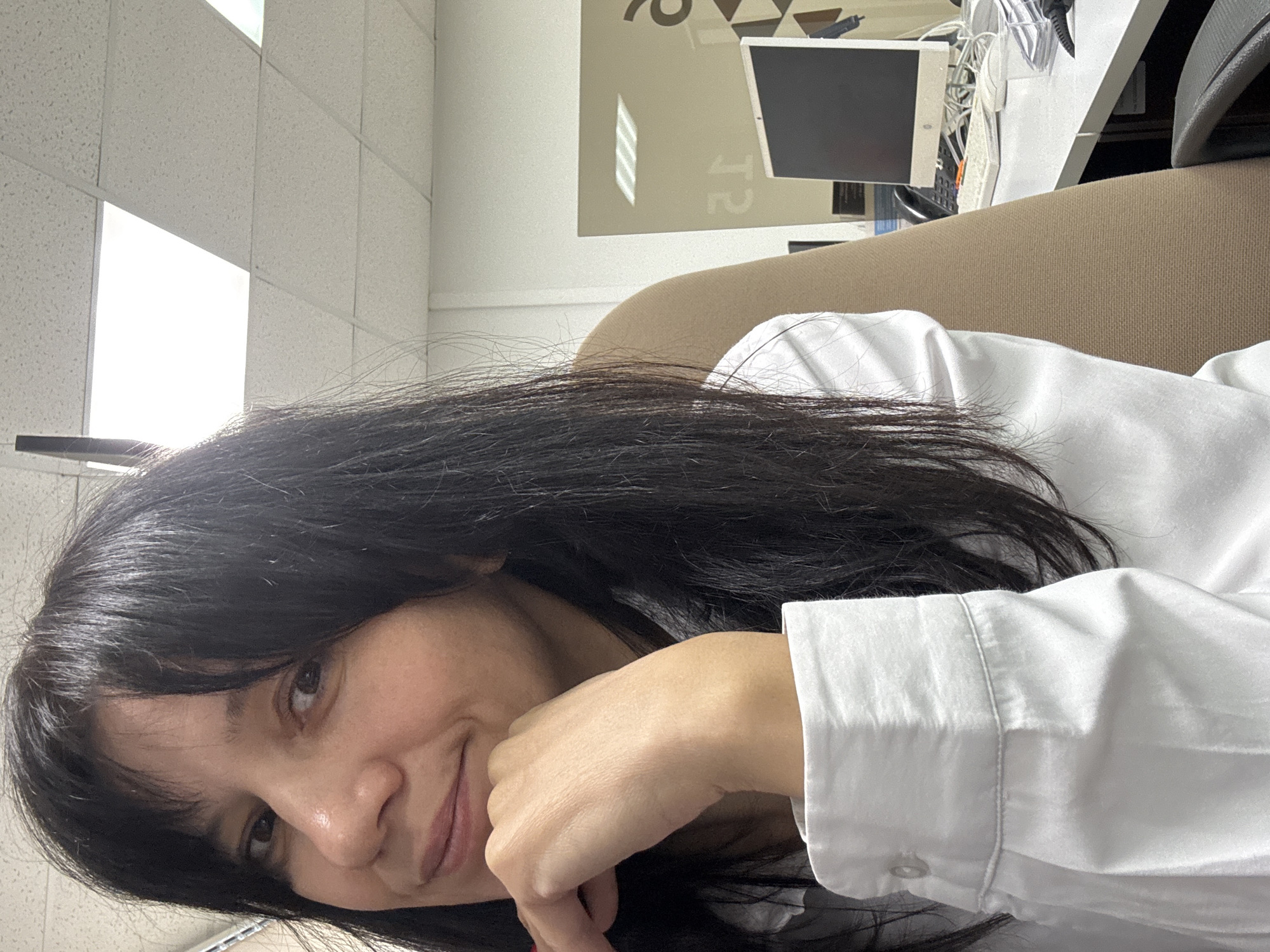 A selfie of a woman with dark hair, wearing a white button-down shirt, resting her chin on her hand. The background includes an office environment with a drop ceiling, a desk with a computer, and a beige chair. It is not possible to determine the location in the city of Chaikovsky, Russia from the image.