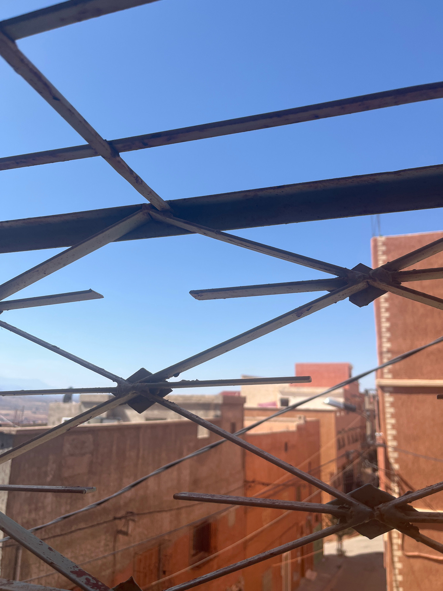 The image captures a view through a weathered metal grate, likely from a window or balcony, onto a sun-drenched city scene. The grate, composed of intersecting metal bars with visible rust and chipping paint, forms a geometric pattern that partially obscures the background. Beyond the grate, a clear, bright blue sky dominates the upper portion of the frame. Below the sky, the characteristic terracotta-colored buildings of Azilal, Morocco, are visible. These buildings exhibit flat roofs and feature small windows and architectural details typical of the region. The urban landscape appears densely packed, with multiple structures clustered together. The overall impression is one of a warm, possibly arid, urban environment under a cloudless sky, viewed through a rustic, protective barrier.