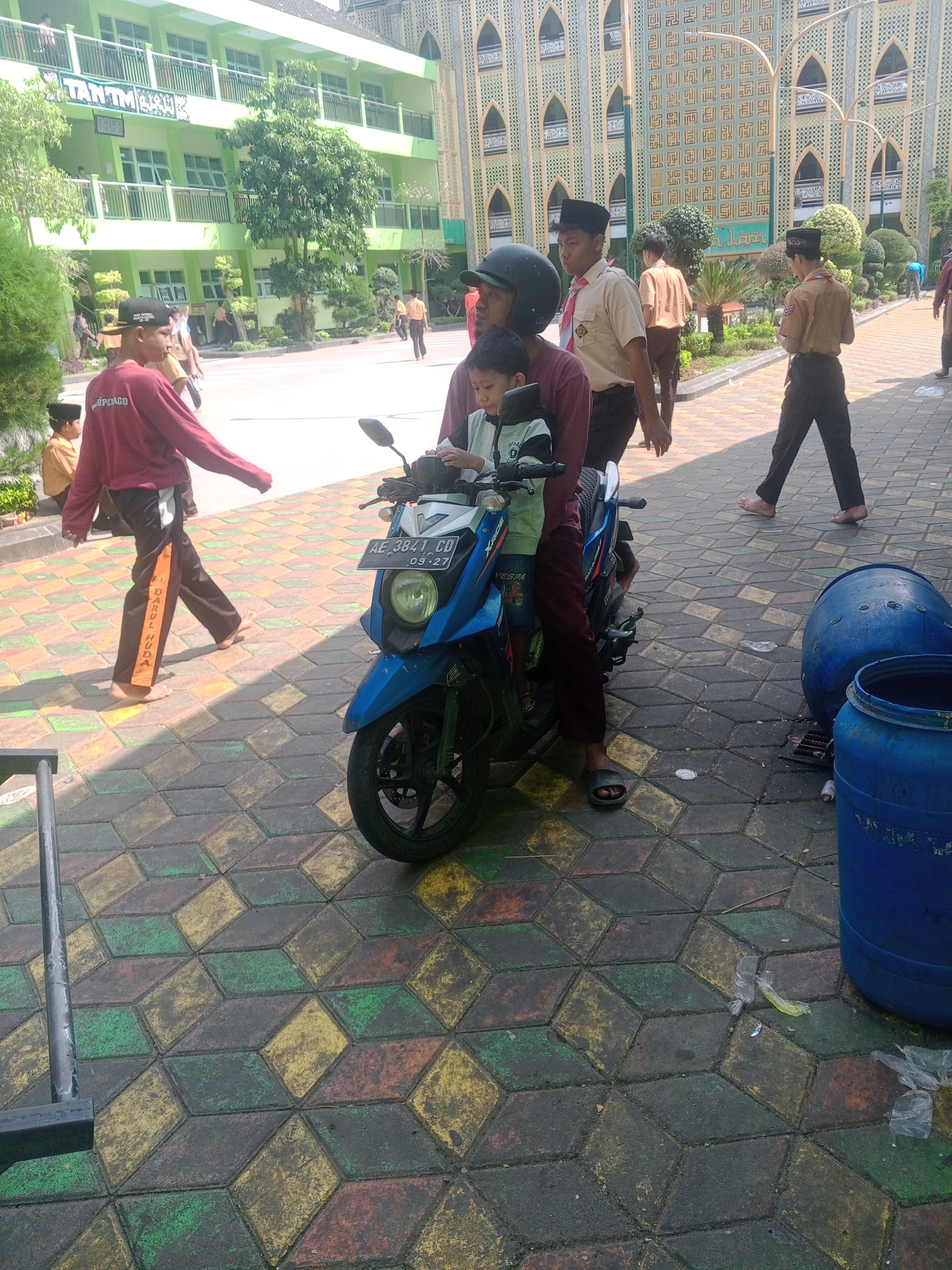 The image depicts a scene in front of a building that appears to be a school or mosque in Ponorogo, Indonesia. A blue motorcycle is parked on a patterned paved ground. A person wearing a maroon shirt and dark pants is seated on the motorcycle, holding a child. The child is wearing a light-colored shirt. Another person, possibly an adult, is standing behind them. In the background, several young people wearing uniforms are walking. One individual in the foreground on the left wears a maroon shirt and dark pants with an orange stripe and the text "DARUL HUDA" written vertically. Two large blue plastic barrels are visible on the right side of the image, near the edge of the paved area. The building in the background is light green with multiple windows and balconies, and features elaborate geometric patterns on its facade, characteristic of Islamic architecture. Trees and greenery are also present around the building. The overall lighting suggests it is daytime.