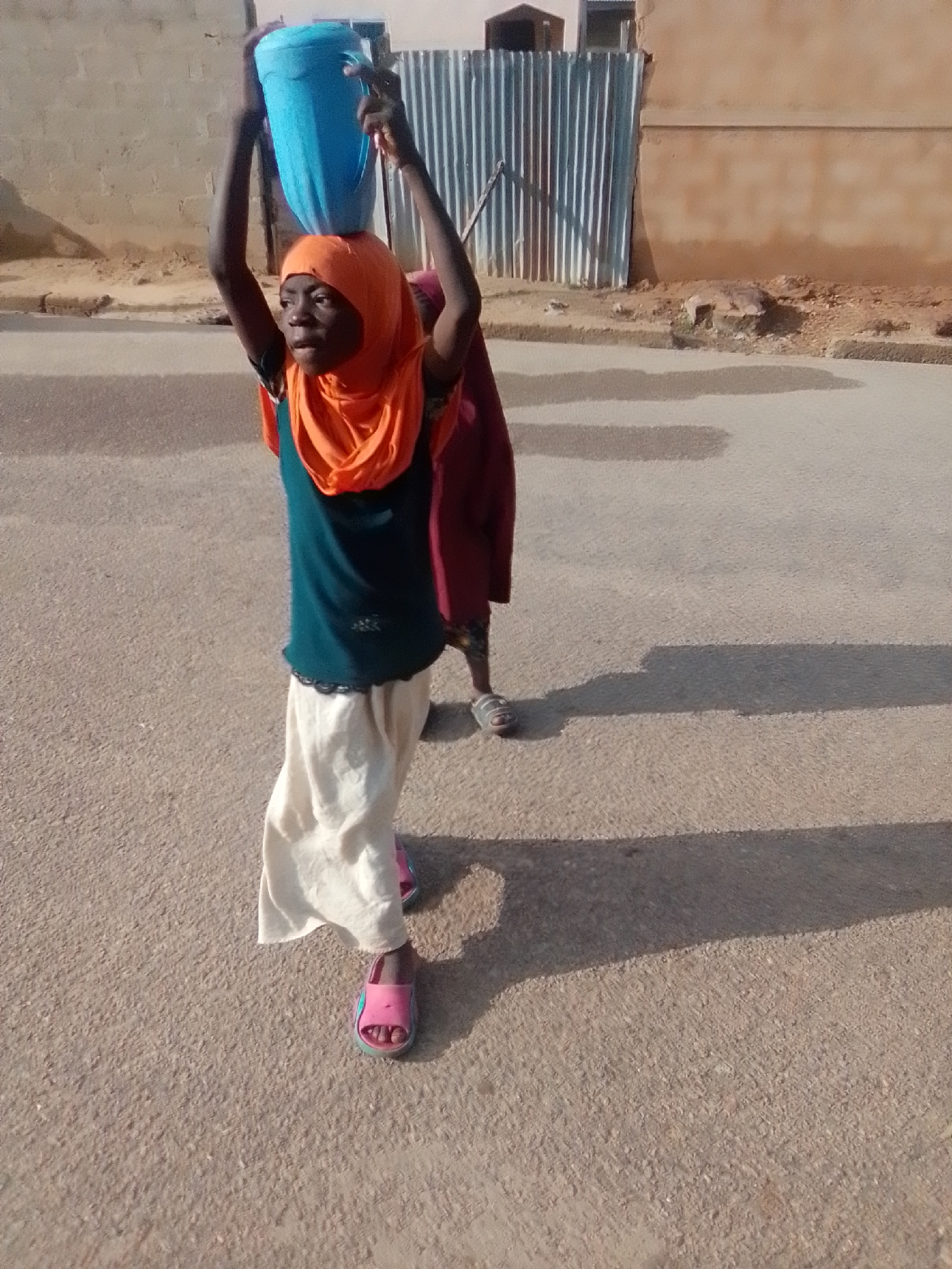 A young person wearing a teal shirt, an orange headscarf, and a cream-colored skirt carries a large, blue plastic bucket on their head. They are walking on a paved road, casting a shadow to their right. Another person, wearing a dark maroon garment, is partially visible behind and to the right of the first person. The background shows a light-colored building with a corrugated metal fence. The scene appears to be an outdoor, daytime setting in Liji Hill, Nigeria.