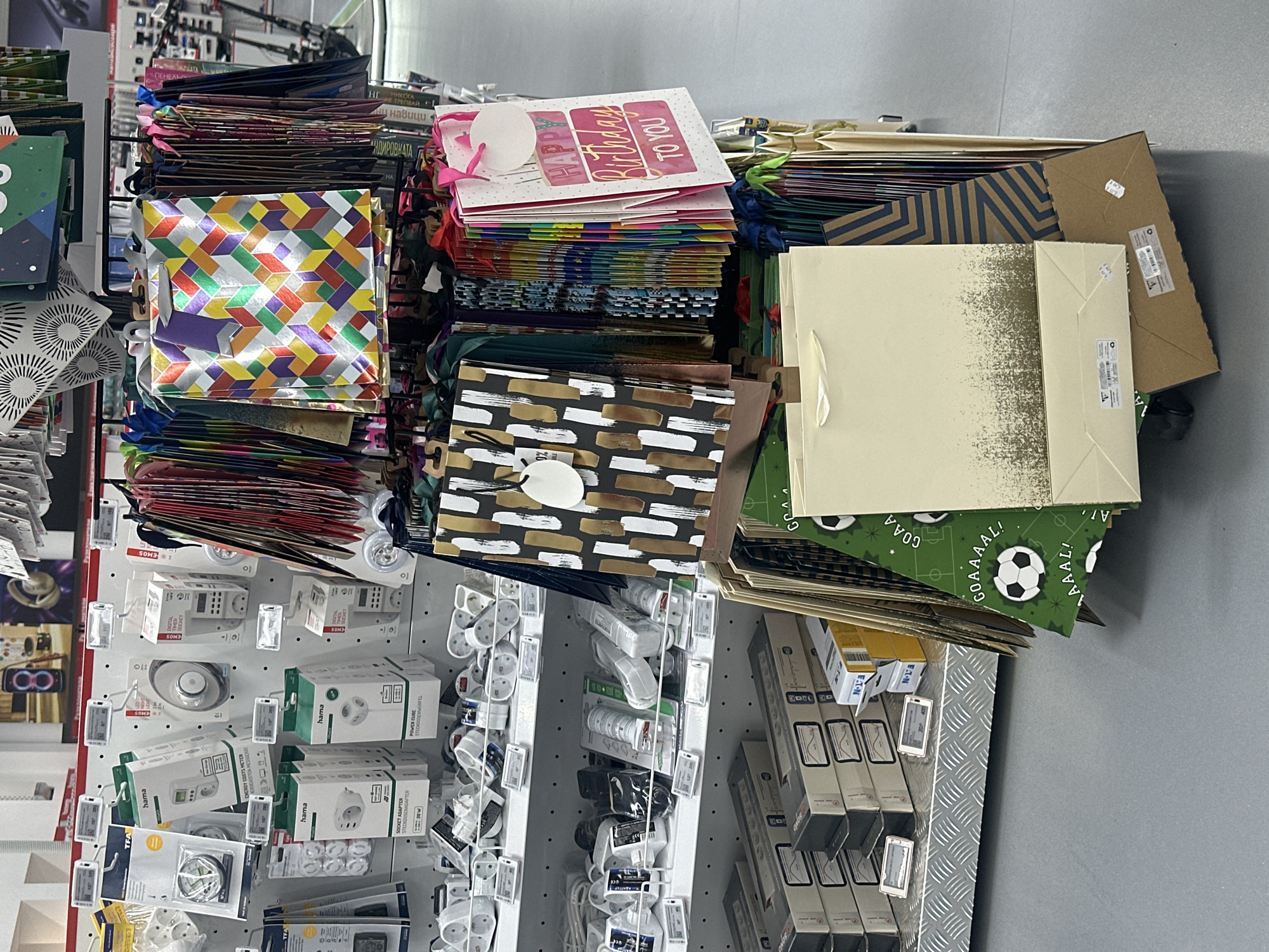 The image shows a shelf in a store, likely in Lovech, Bulgaria, filled with various merchandise. On the left side, there are rows of electronic components and small devices, packaged and displayed on hooks. Labels like "hama" and "EMOS" are visible on the products. To the right, there's a large assortment of gift bags stacked on top of each other. The bags feature a variety of colors, patterns, and themes, including birthday greetings, geometric designs, and sports motifs (soccer). The background includes parts of the store shelving and a glimpse of the floor.
