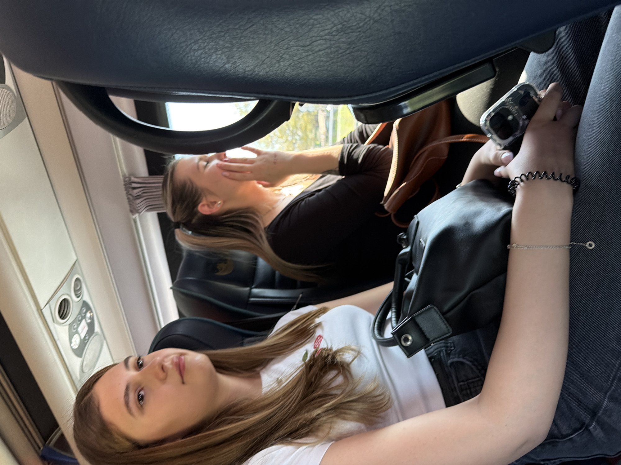 Two women are sitting in seats with dark-colored upholstery. The woman in the foreground is facing the camera, wearing a white shirt with a floral design and black shorts. She has long brown hair. Her left arm is resting on the seat, and she has two bracelets on her left wrist. A black purse is on the seat next to her. The woman in the background is wearing a black top and has long blonde hair. She is covering her face with her hands. A brown bag is next to her. Both women are sitting inside a vehicle, potentially a bus or train, due to the presence of a window and overhead console. The exact location in Plovdiv, Bulgaria, is not discernible from the image.