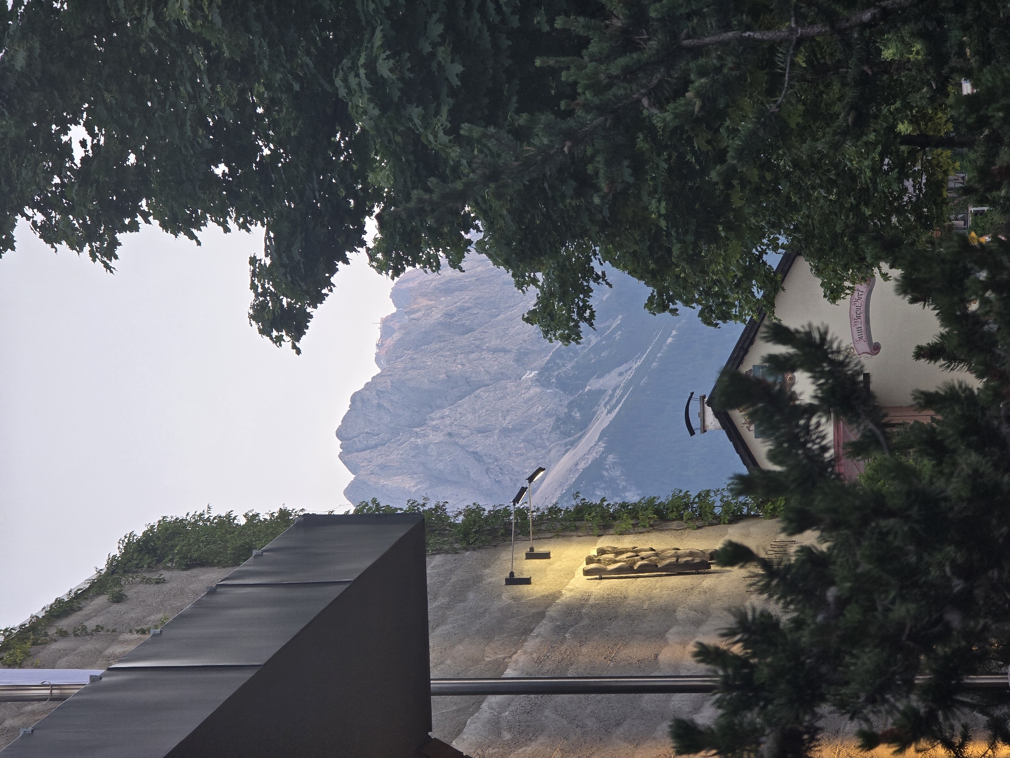 The image depicts a view of the Zugspitze mountain range in Garmisch-Partenkirchen, Germany, framed by architectural elements and foliage. In the foreground, a modern building with dark grey siding and a textured concrete wall is visible on the left. The concrete wall is illuminated by two modern sconce lights casting a warm yellow glow, and a carved relief depicting figures is mounted on it. Lush green vines grow down the wall. To the right, mature trees with dense green foliage partially obscure the view.
In the mid-ground, a cream-colored building with a red-tiled roof is partially visible, featuring a chimney with a curved cap. A decorative pink banner with German text that appears to read "Sinnbieri-Beck" is affixed to the building's facade.
The background is dominated by the imposing, rugged peaks of the Zugspitze mountains, with their steep slopes and rocky formations. The sky above is a pale, hazy blue, suggesting either early morning or late evening light. The overall impression is a serene mountain landscape juxtaposed with modern and traditional architectural elements within a town setting.