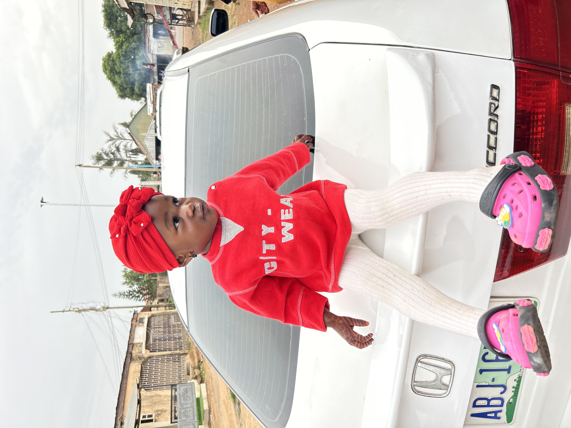 A young child wearing a red headscarf, red sweater with white lettering, and white leggings lies on the back of a white Honda Accord. The child's feet are adorned with pink Crocs with black and floral accents. The car's license plate reads "ABJ-16." The background includes buildings and trees, indicating an urban environment, potentially within the city of Jos, Nigeria. Smoke is visible in the distance. The car's rear window has horizontal lines, and the word "ACCORD" is visible on the right.