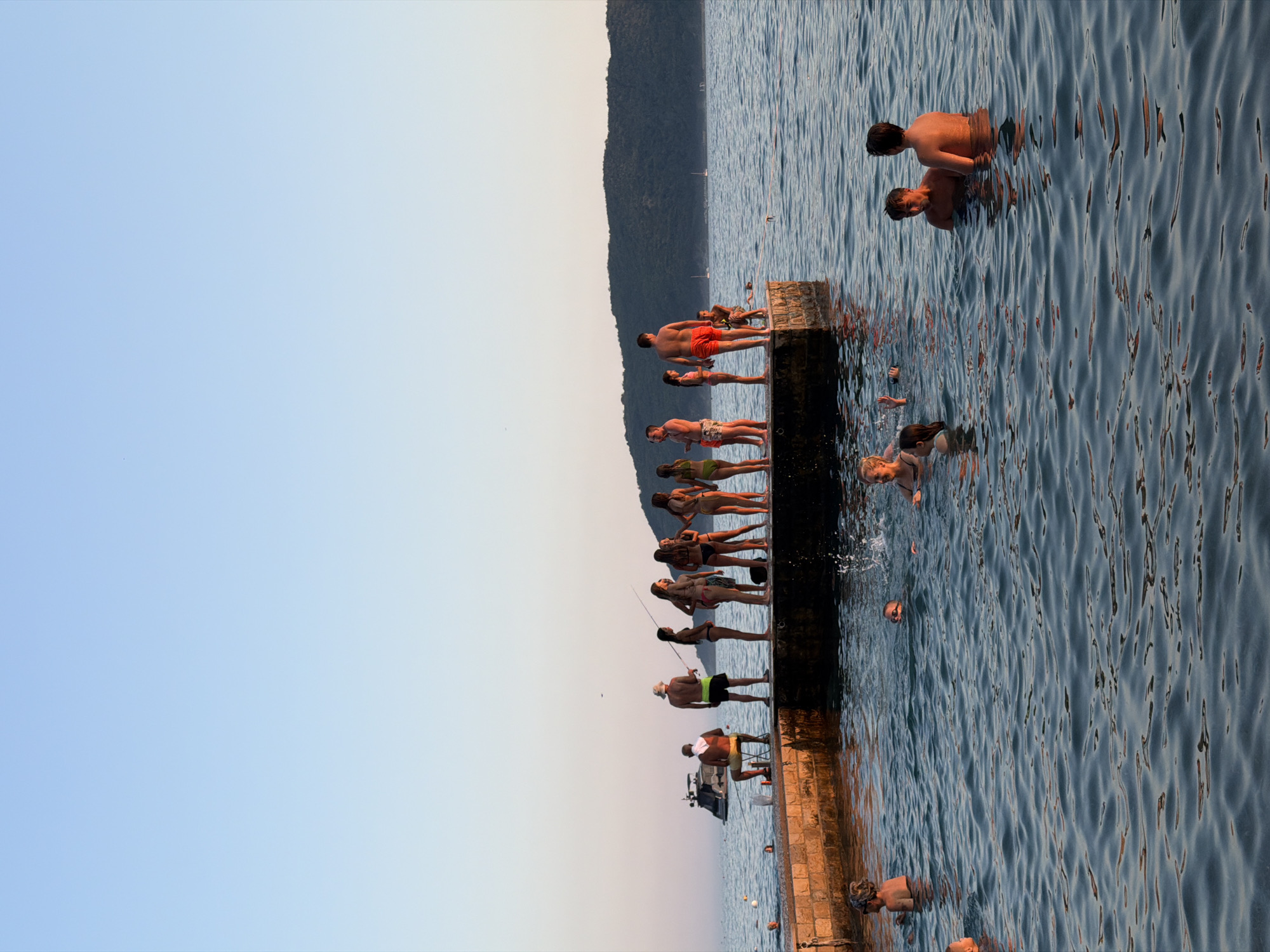 This is a medium-shot, eye-level, full-color photograph taken in Boreti, Montenegro, on what appears to be a clear, calm evening. The scene depicts a group of people enjoying time at a waterfront.
In the foreground, several individuals are in the water. To the left, a person with a light-colored headscarf is partially submerged, looking towards the left. Near the center, a child with wet hair and goggles is visible, and to their right, a woman with blonde hair is smiling and waving. Further to the right, two young men are standing in the water, facing away from the camera and towards the setting sun.
A stone pier extends into the water from the left side of the frame. Along the edge of this pier, a line of people are standing. From left to right: a man with a straw hat and yellow shorts is sitting on a chair, fishing. Next to him, a man in green shorts is standing. Then, a group of women and girls, all in swimwear, are standing and conversing. One of the women is holding a fishing rod. Further to the right on the pier, a man in red shorts is standing, and another individual is seated.
In the background, a dark green, tree-covered hill rises above the water. A small boat with a dark hull is visible on the water to the left of the pier. The sky is a gradient of pale blue, with a hint of pink near the horizon, suggesting either sunrise or sunset. The water is calm, with ripples and reflections visible, and the overall atmosphere is one of leisure and recreation.