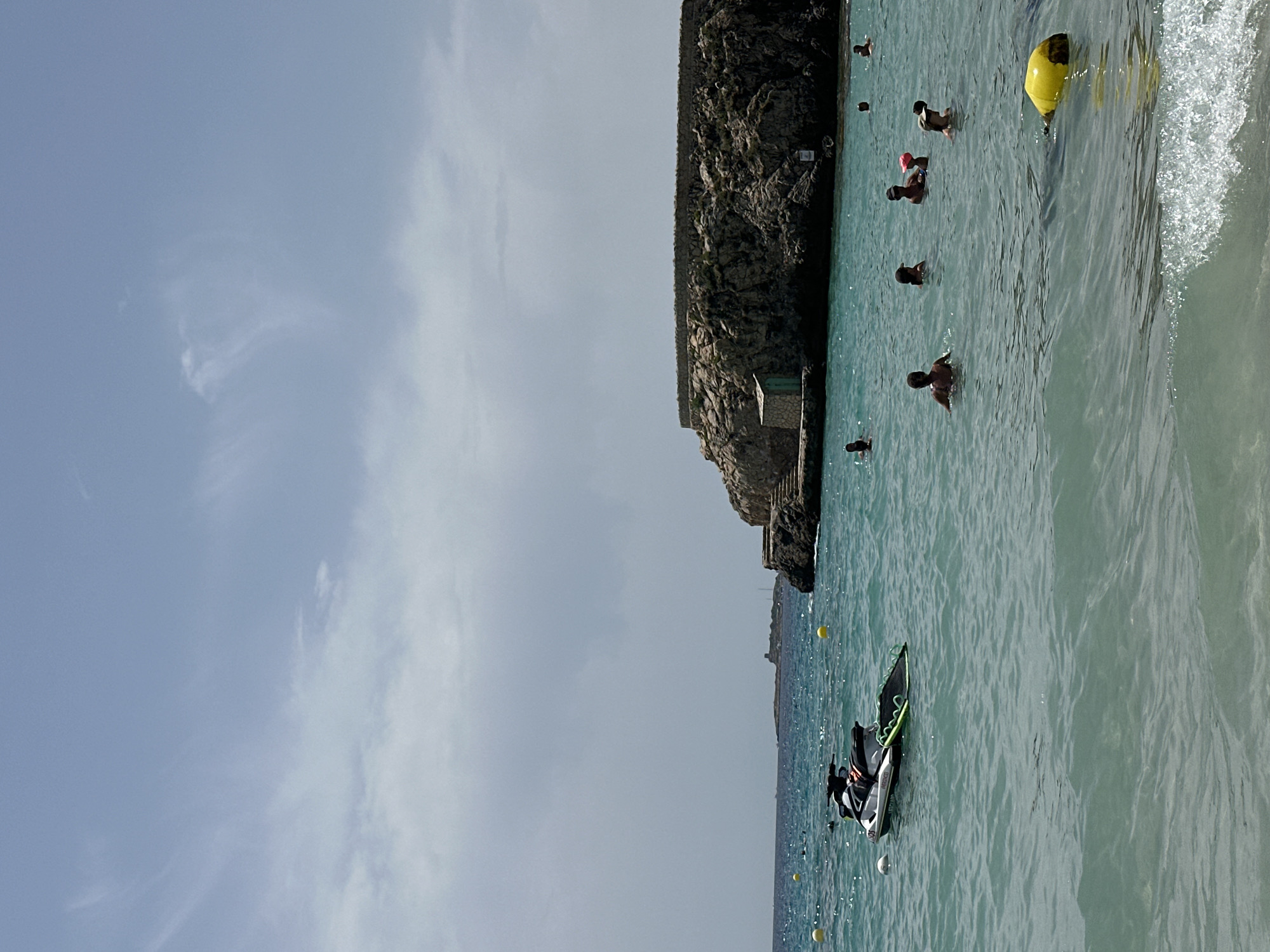 In Sant Agustí, Spain, a jet ski is moored in the turquoise waters of a bay. In the background, a rocky outcrop rises from the sea, topped with a stone-lined circular platform and a small, weathered building. Several people are visible swimming and wading in the shallow water near the shore. The sky is partly cloudy with a pale blue hue. Yellow buoys mark the perimeter of the swimming area. A gentle wave breaks in the foreground, indicating calm seas.