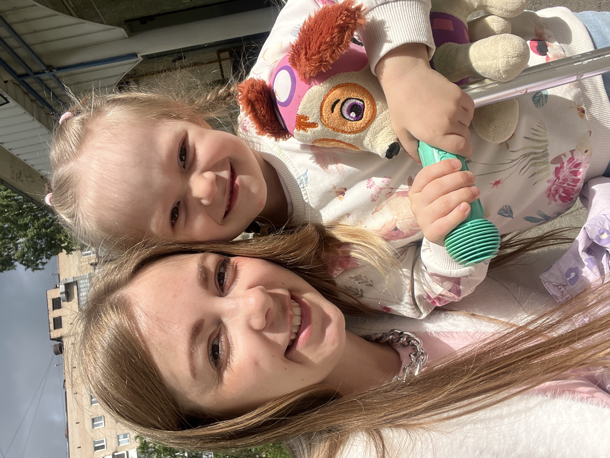 A close-up outdoor portrait captures a smiling adult woman and a young child, likely a mother and daughter, posed in a selfie-style shot. Both individuals have light skin and light hair, and are looking directly at the camera with joyful expressions. The woman, with long light brown hair flowing over her shoulders, is positioned in the foreground, wearing a light-colored top and a silver-colored chain necklace. Slightly behind and above her is the child, who has blonde hair styled in two small ponytails and is wearing a white long-sleeved top adorned with a subtle floral pattern. The child is playfully holding a plush toy that resembles Skye from Paw Patrol, identifiable by its pink aviator hat, and a teal-green toy microphone in her hands. A metallic bar, possibly from a stroller or railing, runs horizontally between the two subjects. The background reveals an overcast sky above residential buildings and some greenery, indicating an urban or suburban outdoor setting. The overall atmosphere conveyed by their broad smiles is cheerful and affectionate. There is no visible text in the image.