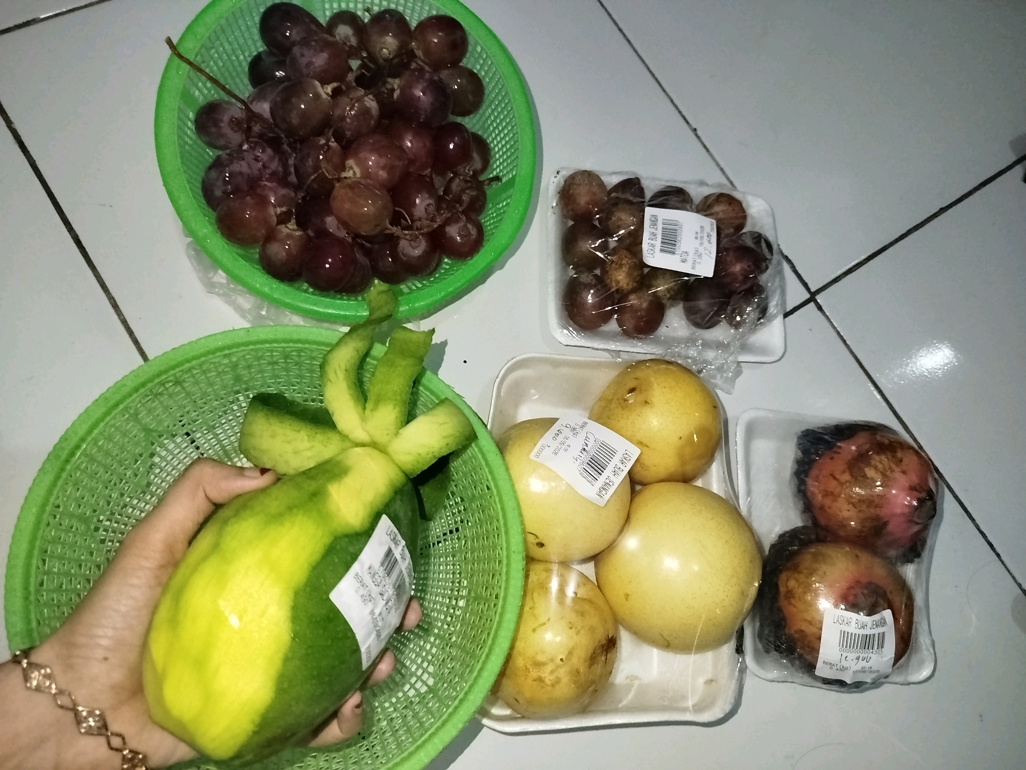 In the city of Babadan, Indonesia, a visually abundant display of fruits is presented on a white tiled floor, illuminated by an unseen light source. In the upper left quadrant, a vibrant green plastic basket overflows with dark purple grapes, their surfaces glistening as if recently washed. To the right of the grapes, a clear plastic-wrapped container holds several small, dark brown fruits, possibly dates or prunes, each exhibiting a wrinkled texture. Adjacent to this container, a white styrofoam tray contains four pale yellow, round fruits, likely pears, also encased in clear plastic. Directly below the pears, another white styrofoam tray is home to two round, reddish-brown fruits, perhaps pomegranates or guavas, also shrink-wrapped. In the lower left quadrant, a large, partially peeled green and yellow fruit, likely a papaya, is held by a person's hand, which is adorned with a delicate gold bracelet. This fruit is positioned within a green plastic basket, similar to the one holding the grapes. The overall scene suggests a grocery shopping or food preparation context, highlighting the fresh produce available.