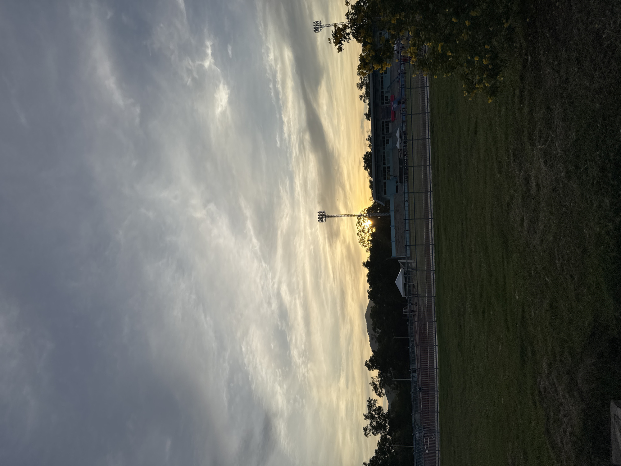 The image depicts an outdoor scene at dusk, likely in a stadium or athletic field. The sky is filled with a mix of dark and light clouds, with the sun partially visible behind a mountain range in the distance, casting a warm glow. In the foreground, a large expanse of green grass covers the ground. Behind the grass, a fence separates it from a track and field area, with stadium seating visible beyond that. Two tall light poles, presumably for illuminating the field, stand on either side of the stadium. Trees line the horizon, and a few tents are set up near the stadium. The overall impression is one of a peaceful twilight scene at a sporting venue.