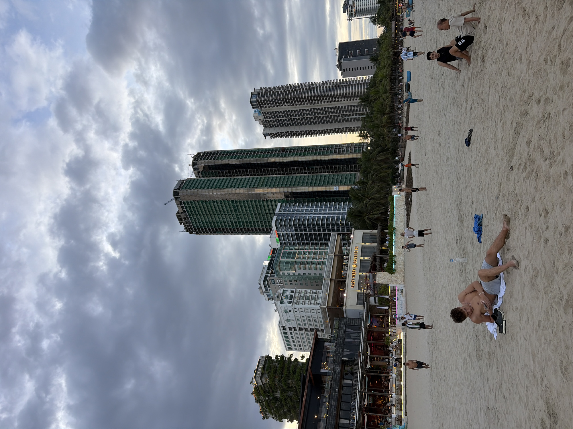 A wide shot captures a cloudy, late afternoon sky over a beach in Đà Nẵng, Vietnam. In the background, a dense cluster of modern high-rise buildings, some appearing under construction, dominate the skyline. The buildings feature a mix of architectural styles, with some clad in glass and others covered in scaffolding and green protective sheeting. A lush, green-covered building, possibly a hotel or apartment complex, stands out on the left side.
In the mid-ground, a strip of restaurants and shops faces the beach, illuminated by warm interior lights. Palm trees line the edge of the development. The beach itself is sandy and populated by a scattering of people.
In the foreground, a young man is lying on a towel, looking at his phone. Another person sits on the sand nearby. Further down the beach, groups of people are engaged in various activities, including what appears to be a game of football or soccer, with children and adults interacting. The overall scene depicts a bustling urban beach environment as daylight begins to fade.