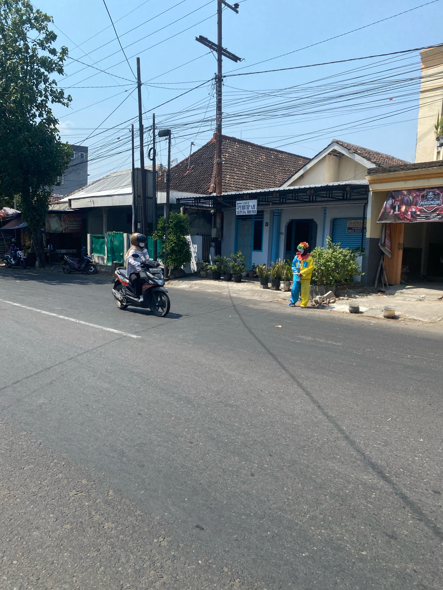 A daytime, eye-level outdoor shot captures a street scene in the city of Kediri, Indonesia. A person wearing a helmet and a striped long-sleeved shirt rides a black and orange motorcycle in the foreground, heading towards the left of the frame. The motorcycle has a license plate. The rider is looking forward, and their face is covered by a mask.
In the background, a light blue building with a white awning stands on the right side of the street. Two individuals dressed in full-body clown costumes, one in blue and yellow and the other in yellow and blue, are standing near the building. One clown holds a red, yellow, and green wig. A green bush is situated between them.
Further to the left, other buildings are visible, including one with a tiled roof. A large tree with green foliage is on the far left. Overhead, numerous electrical wires crisscross the sky, supported by wooden utility poles. The road surface is asphalt, with a white line marking the lane. The weather appears to be clear and sunny.