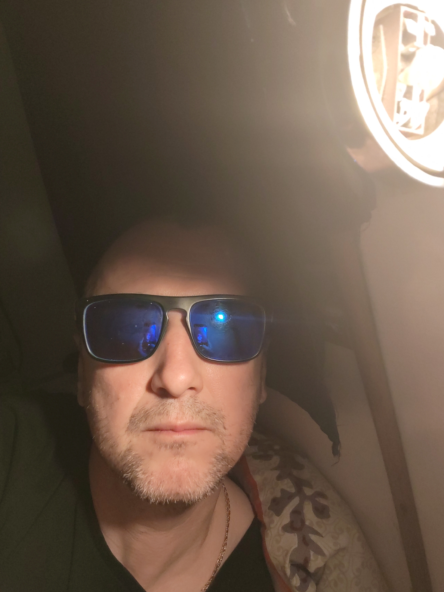 This is a close-up, dimly lit selfie of a man, likely taken indoors in Shcheleiki, Russia. The man appears to be middle-aged, with a bald or closely-shaven head and light stubble or a short beard on his chin and jawline. He is wearing dark, rectangular sunglasses with highly reflective blue lenses, which obscure his eyes. A gold chain is visible around his neck, resting on a dark-colored shirt or garment, possibly green.
A very bright, circular light fixture is prominently featured in the upper right corner of the frame, casting a strong, warm light that illuminates part of the scene and creates intense reflections in the man's sunglasses. These reflections show bright white and blue light sources. The background on the left is dark and indistinct, while the right side reveals a lighter wall or surface and a patterned fabric, possibly a pillow or blanket with a floral or abstract design in brown and cream tones.
The man is looking directly at the camera with a neutral expression. The strong artificial lighting and dark surroundings suggest the photograph was taken at night or in a poorly lit interior space, such as a room, cabin, or vehicle. There is no visible text in the image.