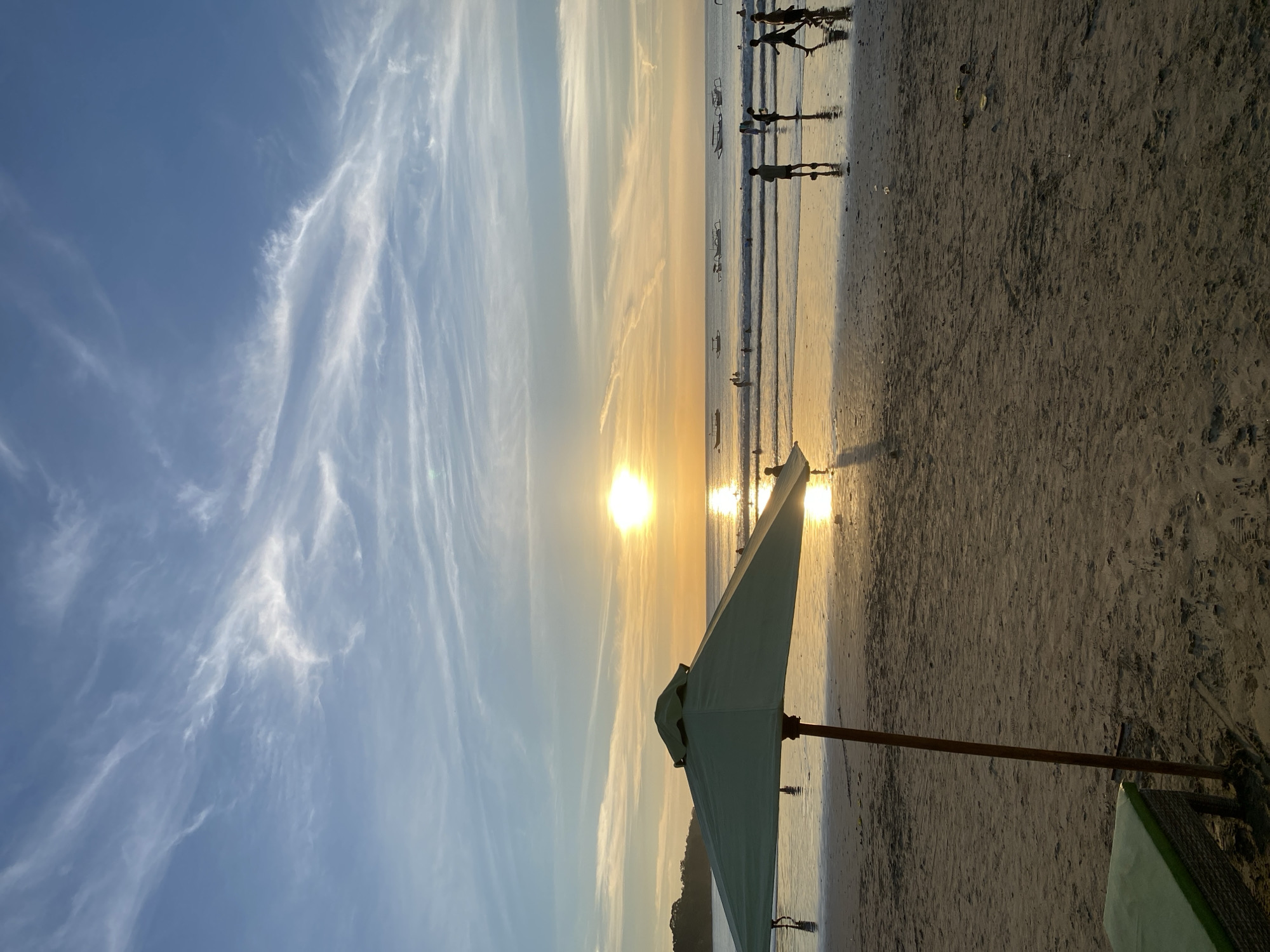 The image shows a sunset scene at a beach in Kuta Selatan, Indonesia. The sun is setting over the ocean, creating a bright reflection on the wet sand. The sky has streaks of cirrus clouds. A beach umbrella is situated in the foreground. In the background, there are silhouetted figures of people near the water's edge, and boats are visible on the water.