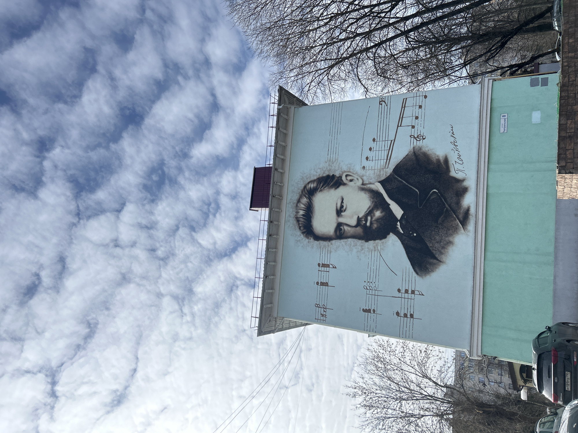 A mural in Klin, Russia, shows Pyotr Ilyich Tchaikovsky's portrait overlaid on musical notation on a building’s light blue wall. The wall is bordered by a light green section. Above the mural, tree branches are visible against a cloudy sky. A building sign is visible on the upper right section of the mural wall. Vehicles are parked in front of the building.