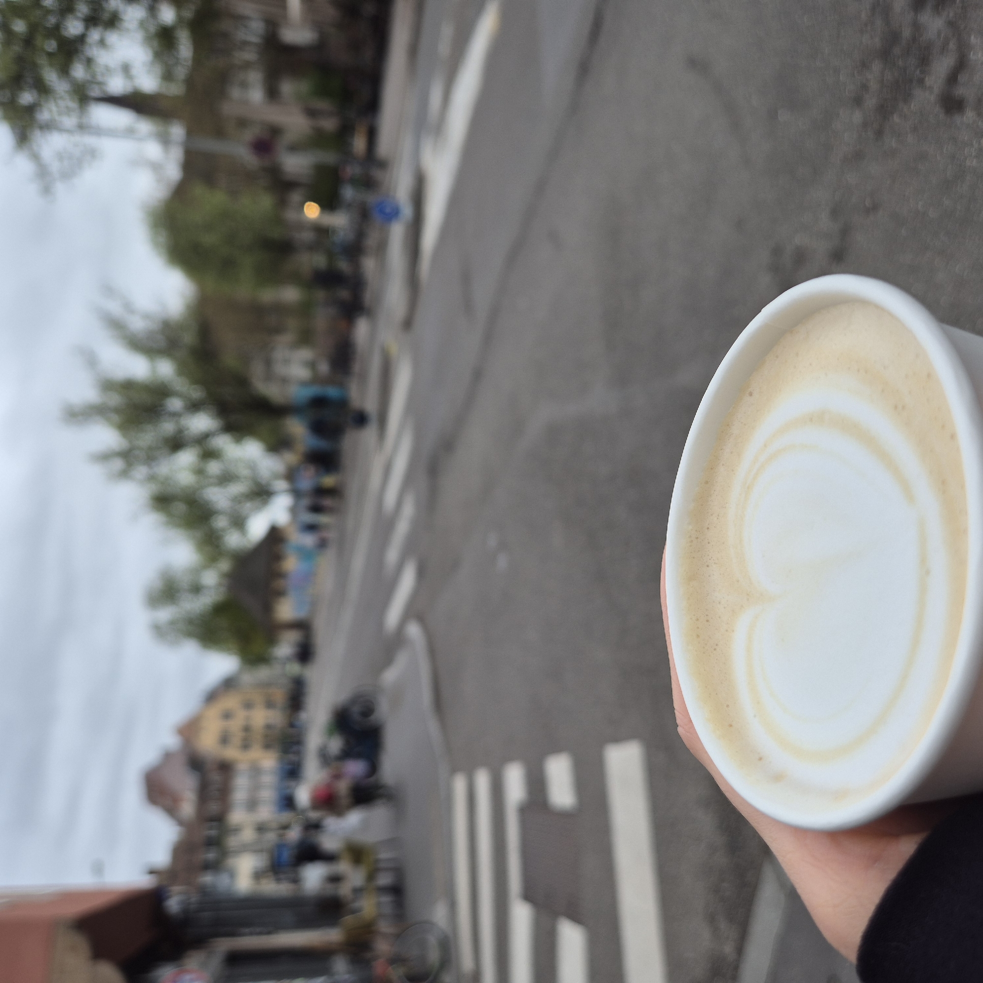 A street-level, eye-level view shows a hand holding a cup of coffee with latte art in the foreground, with a blurred street scene in the background. The location is in Strasbourg, France. The street features a zebra crossing with white stripes, and blurred buildings and trees line the street. A few blurred figures are visible on the crosswalk and alongside the street. The weather appears overcast.