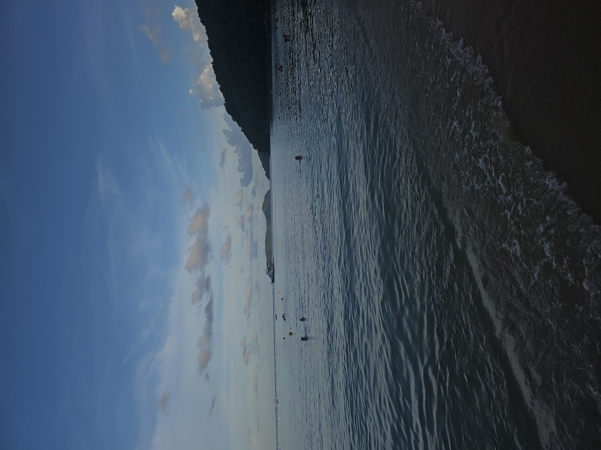 This image depicts a tranquil beach scene in Wichit, Thailand. The sky is a deep blue, with scattered clouds, and the sun appears to be setting or rising, casting a soft light. The ocean is calm, with gentle waves lapping at the shore. Several people are visible in the water, some swimming and others standing. In the distance, a ferry boat is cruising on the water, and a lush green hill forms the backdrop on the right side of the frame. The beach itself is dark sand, with small waves breaking on the shore.
