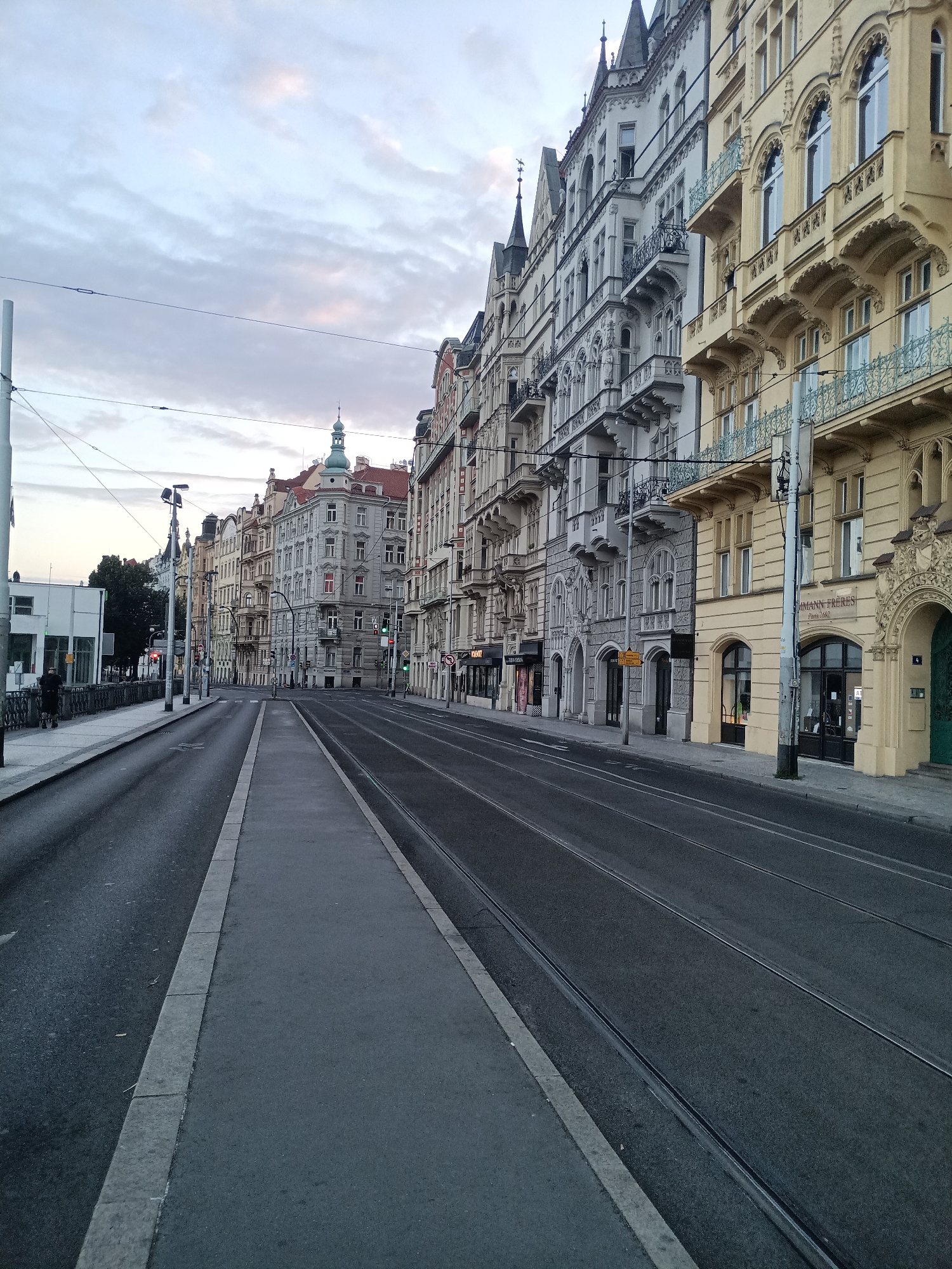 This media file depicts a street scene in Prague, Czechia, likely captured during dawn or dusk. The sky is overcast with subtle hues of pink and blue, indicating the soft light of early morning or late evening. The street features tram tracks, suggesting it's a route for public transportation, and a sidewalk with a person walking on it, implying a quiet, everyday moment.
The dominant subjects are the ornate, historic buildings lining the street. These buildings exhibit intricate architectural details, including elaborate facades, balconies with decorative railings, arched windows, and steeply pitched roofs, characteristic of Prague's historical architecture. Some buildings have a pale yellow or off-white facade, while others are grey. Several shops are visible at street level, with some signage hinting at their businesses.
The scene conveys a sense of calm and stillness, possibly due to the early or late hour. The lighting is subdued, and the weather appears to be mild, with no visible precipitation. The tram tracks and the well-maintained sidewalk and road suggest an urban environment.
Visible text includes a sign that appears to read "SAMANN FRERES Paris 1662," suggesting a historical business or brand. Another sign with red text is partially visible on one of the buildings. Traffic lights are also present, indicating the flow of traffic even in this tranquil setting. The overall impression is that of a serene moment in a historically rich European city.