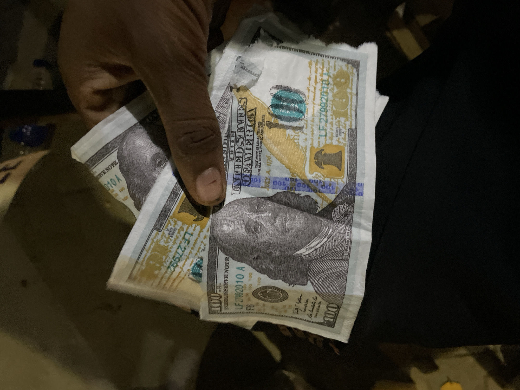 A close-up, overhead shot captures a dark-skinned hand holding several US $100 bills, partially obscuring the face of Benjamin Franklin on the front of the banknotes. The bills, some appearing newer than others, are fanned out, with visible serial numbers such as "LF 27992010 A" and "LF 27992018 A." The background is dark and indistinct, with hints of urban environmental elements suggesting a nighttime outdoor setting. This image does not contain any specific visual cues that would place it within Lekki, Nigeria, or any other identifiable city.