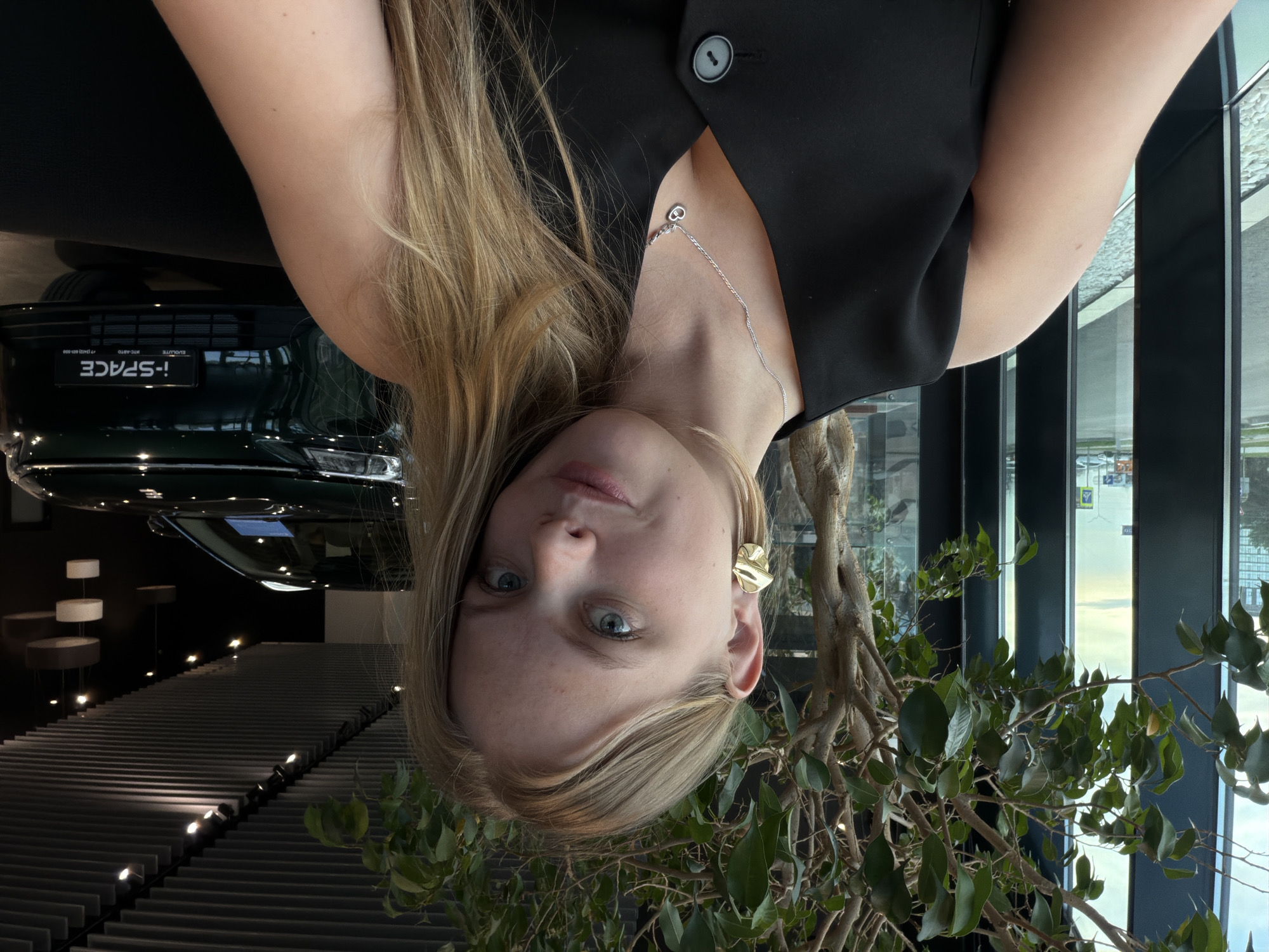 The image is taken indoors. A fair-skinned woman with blonde hair is centrally featured. She wears a black vest over a top, a silver necklace with a small heart pendant, and a gold ear cuff on her left ear.
Behind her to the left, partially visible, is a dark green I-PACE vehicle. The interior includes a set of minimalist white cylindrical displays on dark poles, and a textured surface with rows of recessed lights.
To her right, a large leafy plant is situated in front of a window. The view through the window suggests a building or cityscape. Given the question prompt, the location is potentially in Izhevsk, Russia, but no specific landmarks are evident in the image to confirm.