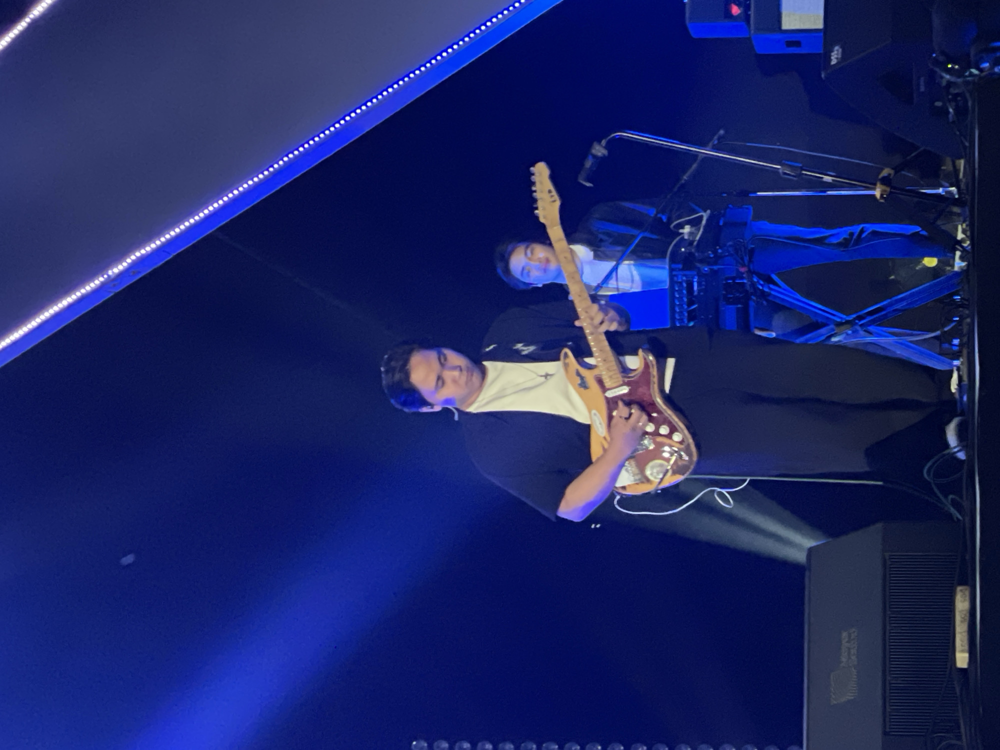 The image captures two individuals performing on a stage illuminated by blue lighting. A man in a black shirt and white t-shirt is playing an electric guitar. Behind him, another man is playing a keyboard. Microphones and speakers are also visible on the stage. The location is Salembaran, Indonesia.