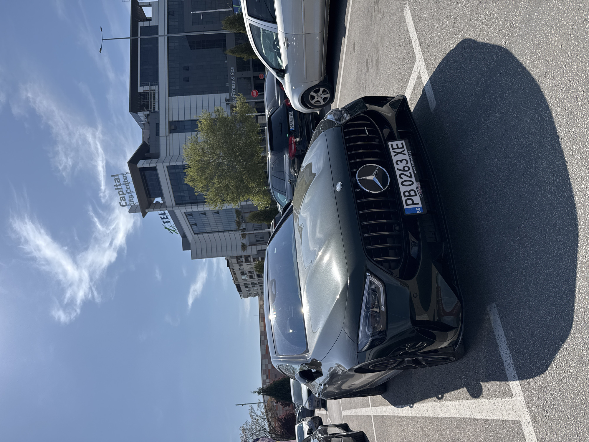 This media file captures an outdoor parking lot scene on a bright, sunny day in Plovdiv, Bulgaria. In the foreground, a dark-colored Mercedes-Benz vehicle, likely a sedan or SUV, is prominently parked, its glossy surface reflecting the sunlight. Its front grille, featuring the Mercedes-Benz logo, and the Bulgarian license plate "PB 0263 XE" are clearly visible. Below the license plate, the text "www.avtosviat.bg" is also legible. Strong shadows are cast on the asphalt, indicating the sun is high in the sky.
Behind the Mercedes, other parked cars are visible, including a silver hatchback and a dark SUV with the Bulgarian license plate "PA 0609 MB". In the background, a modern, multi-story building dominates the scene. Large signs on the building clearly display "Capital City Center" and "TEL", with "Fitness & Spa" also visible on another section. The architecture suggests a contemporary commercial or office complex. The sky is a clear blue with some wispy white clouds. A "STOP" sign is partially visible near some trees in the mid-ground. The overall impression is a vibrant, active urban environment during daylight hours.