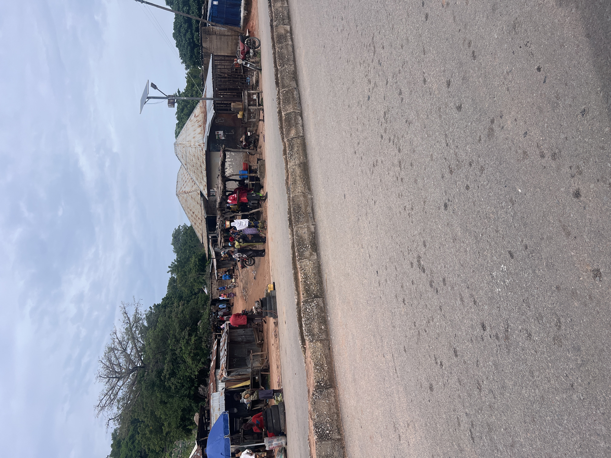 This image depicts a street scene in Kona Garu, Nigeria, likely during daylight hours given the overcast sky and the general lighting. The primary subjects are a collection of makeshift structures that appear to be shops or stalls lining a paved road. These structures are constructed with corrugated metal roofs and wooden frames, suggesting a developing or informal commercial area.
Several people are visible amidst the stalls, engaging in various activities. Some are standing or walking, while others appear to be interacting with vendors or examining goods. There are also several motorcycles parked near the stalls, indicating a common mode of transportation in the area. Some of the stalls display goods for sale, though the specific items are not clearly discernible. One stall has a blue umbrella providing shade. A solar-powered street light is visible, suggesting some modern infrastructure amidst the more traditional setting.
The road itself is asphalt and appears to be in fair condition, with some dark spots suggesting oil or water stains. A concrete curb separates the road from the area where the stalls are situated. Behind the stalls, dense green foliage, including trees, suggests a natural landscape bordering the settlement. The sky is cloudy, indicating overcast weather, which might imply a humid or potentially rainy climate. The overall impression is one of a busy, everyday commercial activity in a Nigerian community. No visible text is present in the image.