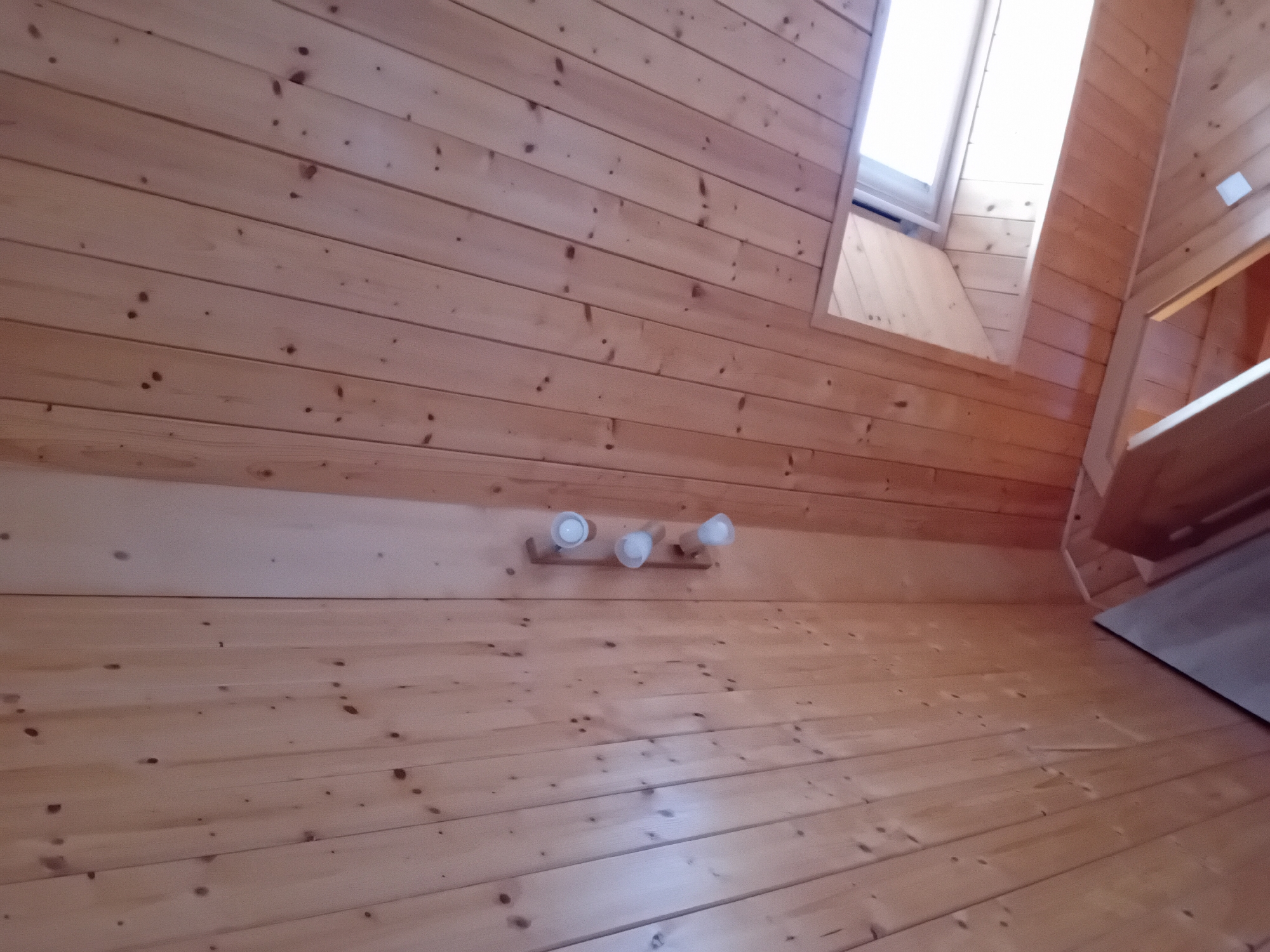 This is a low-angle interior shot of a room with a wooden plank ceiling. The ceiling planks run diagonally from the bottom left to the top right of the image. In the center of the image, a wooden fixture with three white light bulbs is mounted on the ceiling. To the right of the light fixture, a portion of a sloped skylight is visible, letting in bright daylight. The overall impression is one of natural wood construction, likely in a rustic or cabin-like setting. This room is located in Zuberec, Slovakia, a village known for its traditional folk architecture and natural beauty, suggesting this could be a residential interior or a building within an open-air museum like the Zuberec - Brestová Múzeum or a holiday cottage.