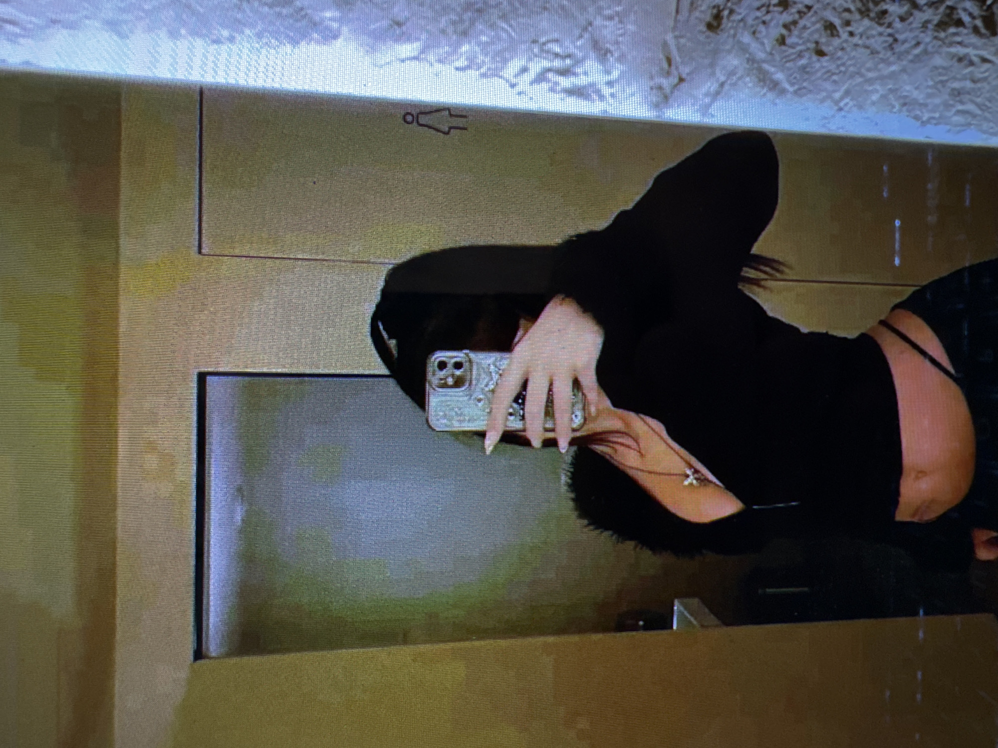 In a dimly lit room, a person is taking a mirror selfie. They are wearing a black, fuzzy-collared, cropped jacket with a zipper, a diamond-shaped pendant necklace, and low-rise pants that reveal a thong. The person is holding a smartphone with a decorative case in their right hand. The background shows a wall with two doors and a sign indicating a women's restroom. The location is not specified as Castelo Branco, Portugal; this detail is not derivable from the image.