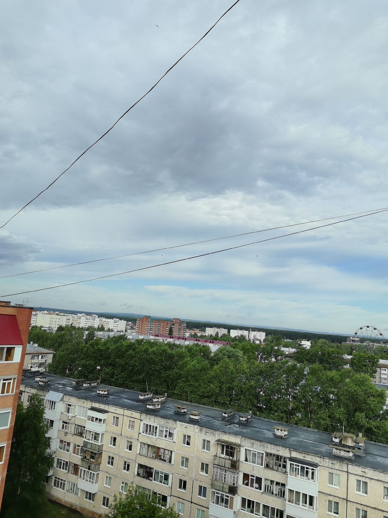 This image provides an aerial view of the city of Neftekamsk, Russia, on an overcast day. The foreground is dominated by the rooftops and upper floors of several Soviet-era apartment buildings, characterized by their pale yellow and grey facades, numerous balconies, and ventilation shafts on the roofs. A brick building with a red roof is partially visible on the left. Behind the apartment buildings, a dense line of green trees stretches across the middle ground, obscuring the lower parts of distant structures.
Further in the background, a cityscape unfolds with a mix of buildings of varying heights and colors, including what appears to be a Ferris wheel on the right side, suggesting an amusement park or recreational area. A few birds are visible in the sky, and power lines crisscross the frame diagonally. The sky is heavily clouded, with patches of lighter blue peeking through in some areas. The overall impression is of a typical urban landscape in a Russian city, likely during the warmer months given the lush greenery.