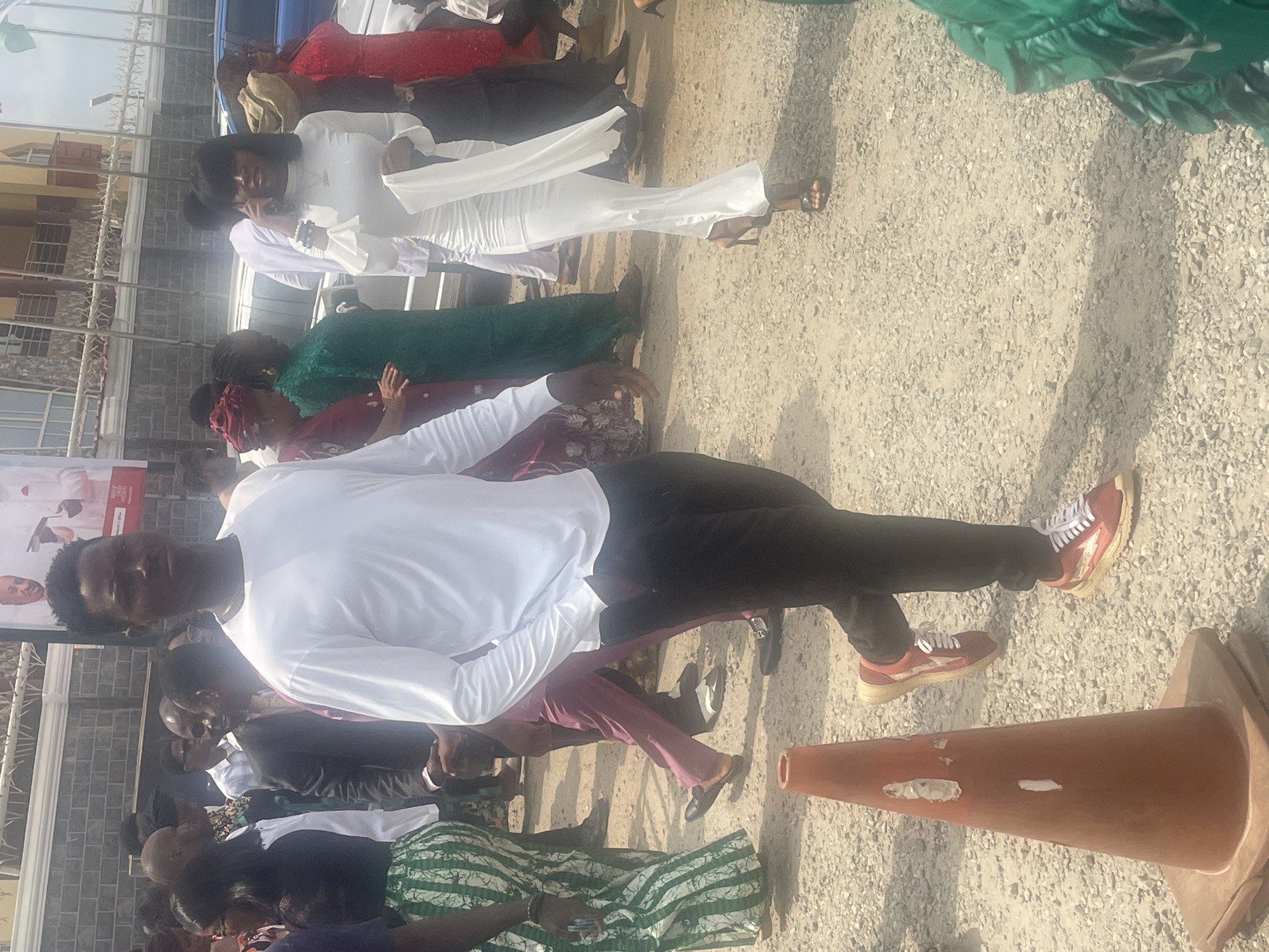 A group of people are standing outside on a gravel lot, likely in Abuja, Nigeria based on context. A man in a white shirt and black pants with red sneakers is sitting near an orange traffic cone. Several women are wearing colorful dresses, and a woman in white is talking on a phone. The scene is outdoors with a building in the background.
