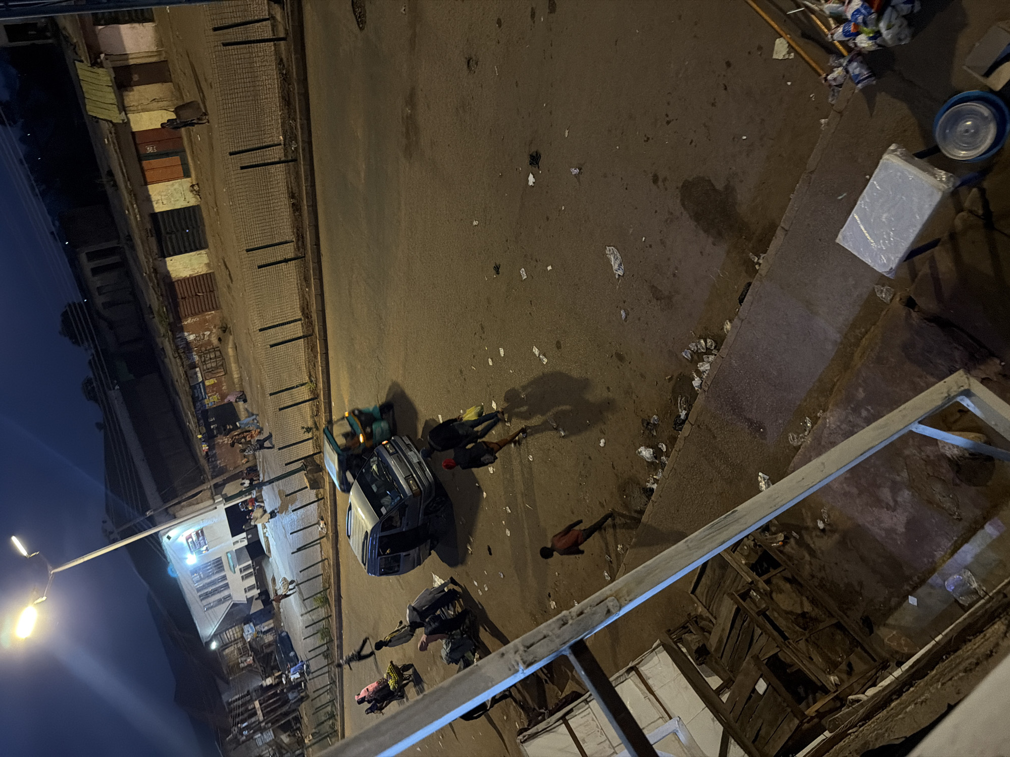 This media file shows a street scene at night in Bukuru, Nigeria. The viewpoint is from above, looking down at the street and sidewalk. Several people are visible on the street, some appearing to be gathered around a parked vehicle. The lighting suggests it is nighttime, with a street lamp casting illumination on the scene. There is scattered debris on the street and sidewalk, including some plastic bags and other refuse. A car is parked on the street, and a smaller vehicle, possibly a tricycle or motorcycle taxi, is also present. The surrounding buildings appear to be commercial or residential, with shutters or doors visible. The overall impression is one of a somewhat disorganized and possibly bustling urban environment at night. There is no discernible text in the image.