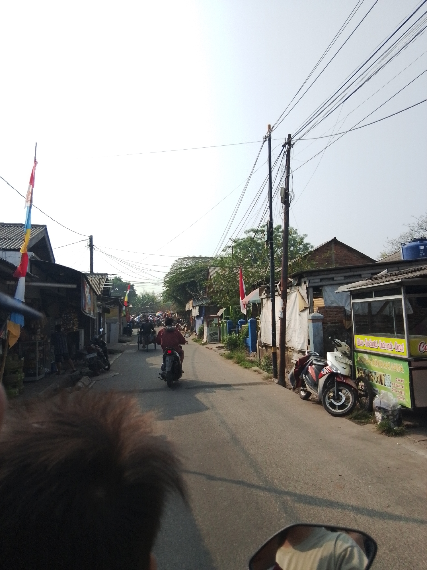 The image depicts a street scene in Bencongan, Indonesia, on a bright, clear day. The street is paved and lined with buildings on both sides, appearing to be a mix of residential and commercial structures. Overhead, a dense network of electrical wires crisscrosses the sky, supported by wooden utility poles.
On the left side of the street, a row of low-rise buildings with corrugated metal roofs and various storefronts is visible. Colorful flags, including what appear to be Indonesian flags, are displayed on poles along this side. Several motorcycles are parked along the sidewalk.
The right side of the street features more buildings, some with brickwork and corrugated roofs, and others with tarpaulin coverings. A food stall with a green and yellow sign is prominent, with a display case showcasing items. A white and red motorcycle is parked in front of the stall.
The street itself is occupied by various forms of transportation. In the foreground, a person's head and shoulder are visible, suggesting the photo is taken from the perspective of someone on a motorcycle. Further down the street, a person is riding a motorcycle, and behind them, a three-wheeled vehicle, possibly a becak or a motorized rickshaw, is also moving. Other motorcycles and people are visible in the distance, indicating moderate street activity. The overall impression is a bustling local street with a vibrant atmosphere.