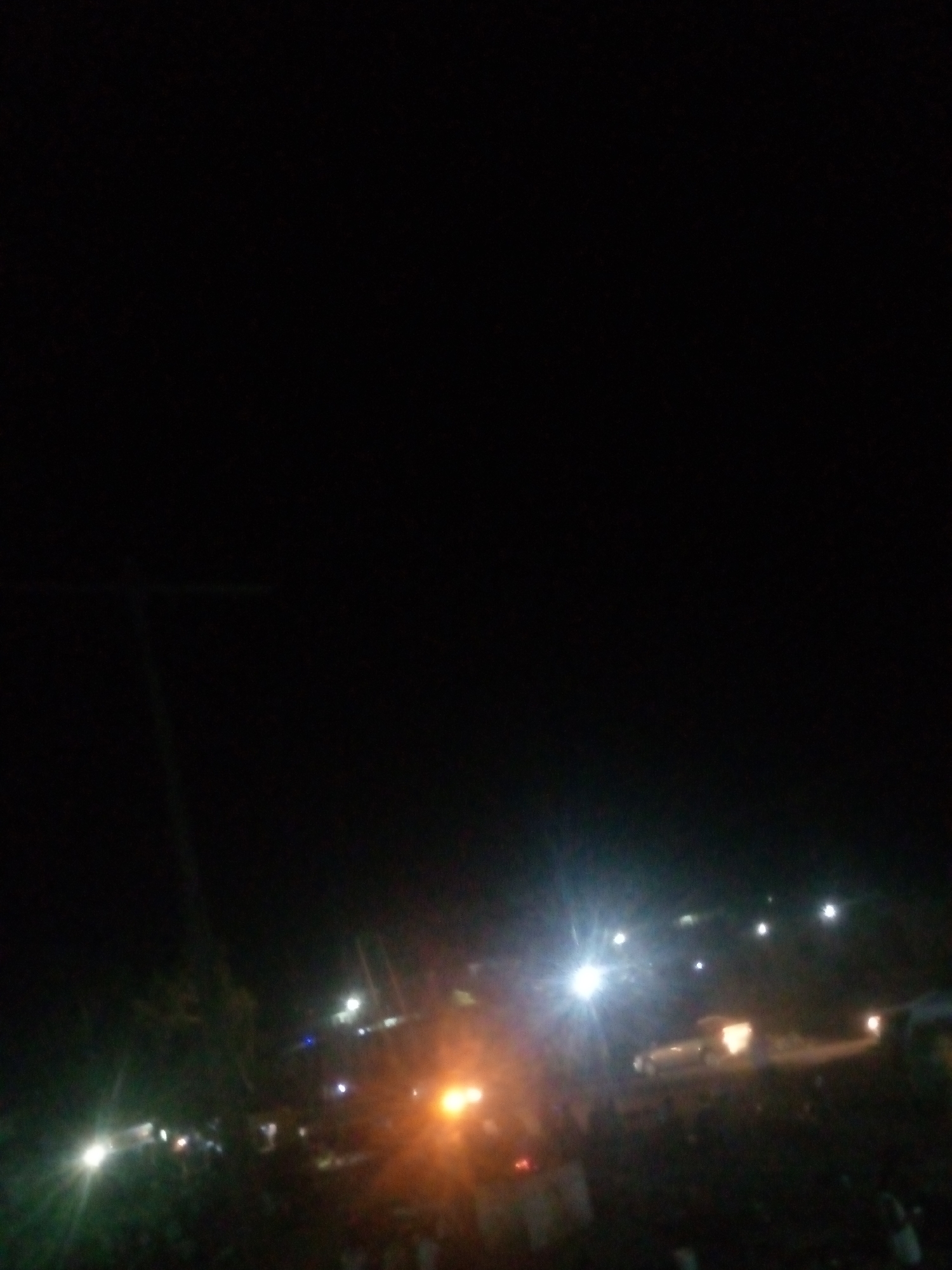 The image is a low-light, blurry photograph taken at night in Jalingo, Nigeria. The scene appears to be an outdoor gathering with multiple artificial light sources. In the foreground, a dense crowd of people is visible, though their features are indistinct due to the poor lighting and blurriness. Several bright lights, some appearing to be headlights or streetlights, illuminate parts of the scene, creating lens flares and a hazy effect. In the background, more lights suggest buildings or structures. The overall impression is of a busy, illuminated area within the city at night, possibly a market, street festival, or a gathering of some sort. The terrain is not clearly discernible due to the darkness and blur.