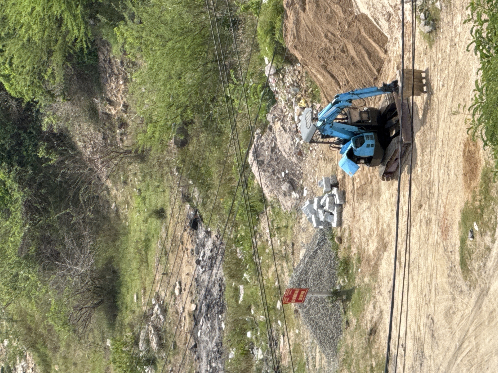 This is a medium-distance, high-angle photograph of a construction site in Bac Nha Trang Ward, Vietnam. A blue excavator is the central subject, positioned on a dirt ground. It is facing right, with its arm extended downwards and a wooden container beneath its bucket. To the left of the excavator, a pile of gravel is visible, with a red sign on a pole in front of it. Adjacent to the gravel pile, to the right, are several stacked blocks of grey stone. A large mound of tan sand is situated to the right of the excavator. In the background, dense green vegetation covers a sloping hill. Several black wires are strung across the scene horizontally. The ground is uneven and appears to be a mix of dirt and scattered rocks.