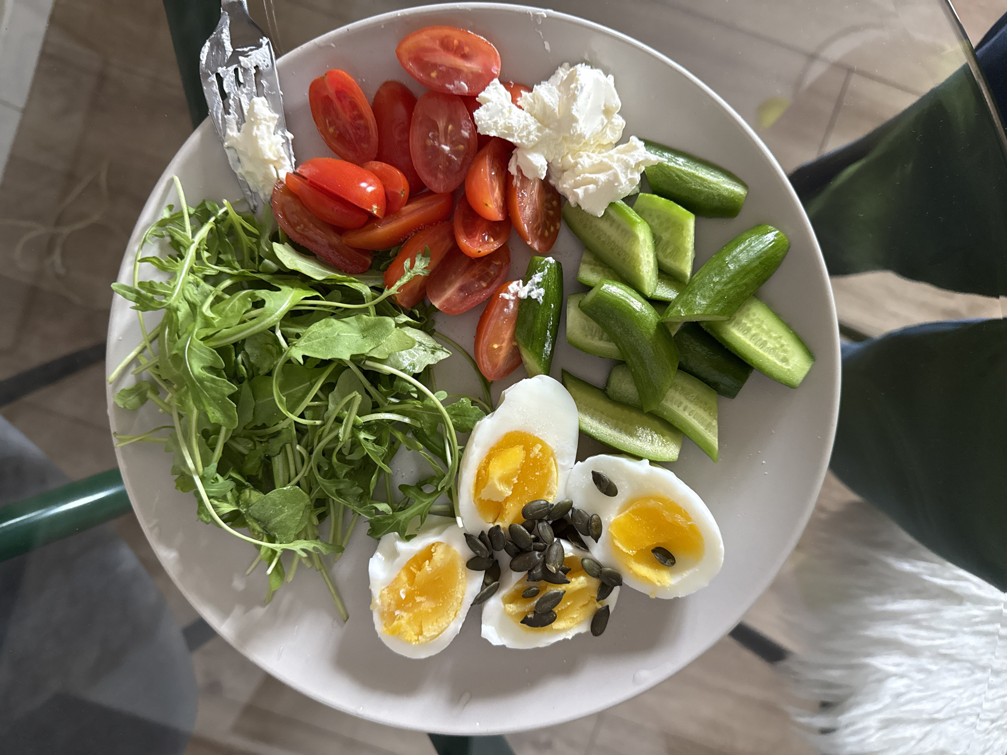 A top-down shot depicts a white plate filled with a salad comprised of various components. There is a large pile of arugula, a serving of sliced cherry tomatoes, a dollop of a white, soft cheese, pieces of sliced cucumbers, and three halved hard-boiled eggs sprinkled with dark seeds. A fork rests on the side of the plate.
The plate sits on a glass table. Part of a green chair frame is visible in the lower left corner, while a white, fluffy object is seen in the lower right.
The location is unidentifiable as Tel-Aviv based on the information in the image. No landmark or cultural object indicative of Tel-Aviv is present.