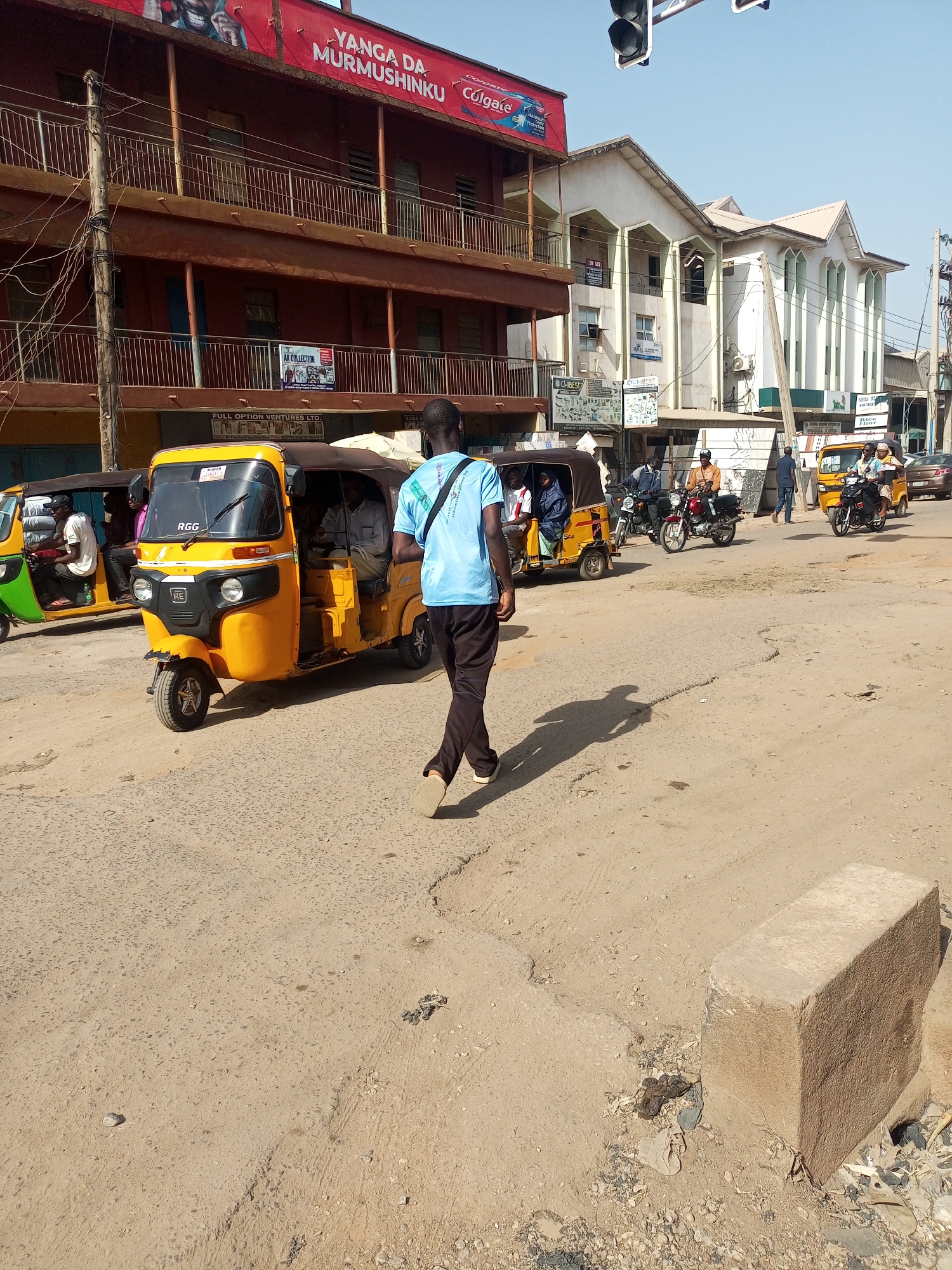 The image depicts a street scene in Maiburiji, Nigeria. A man wearing a blue shirt and dark pants walks across a dirt road, away from the camera, casting a shadow. Several auto-rickshaws, also known as "keke" or "tricycles", are present, some carrying passengers. In the background, there are several motorcyclists. The buildings that frame the background feature diverse architectural styles and colors, including a red multi-story structure with a "YANGA DA MURMUSHINKU Colgate" advertisement at the top, and a lighter-colored building with multiple arched windows. Other visible signs are "AK COLLECTION" and "FULL OPTION VENTURES LTD", "CHIBEST". The road itself is unpaved and appears worn. In the foreground on the right, a rectangular concrete block sits at the roadside. Traffic lights are visible in the top portion of the image.