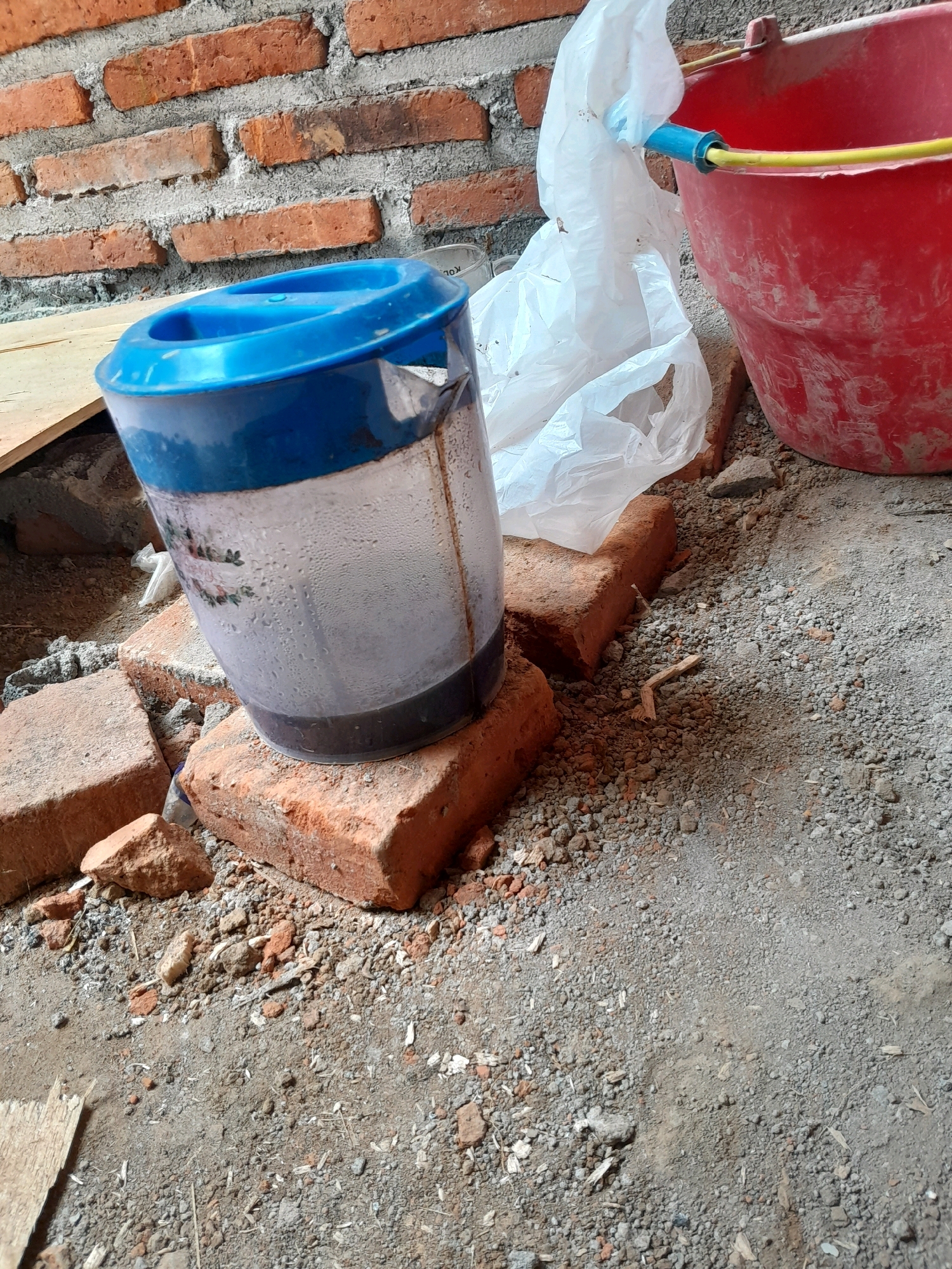 The image depicts a construction or renovation site. A brick wall is visible in the background, along with scattered bricks, gravel, and dirt on the ground. A red bucket with a yellow hose is positioned to the right, and a plastic bag is draped over some of the bricks. In the foreground, a translucent jug with a blue lid sits on a brick. The jug contains a dark liquid and has condensation on its exterior. The scene suggests ongoing construction work in Karangduren, Indonesia.