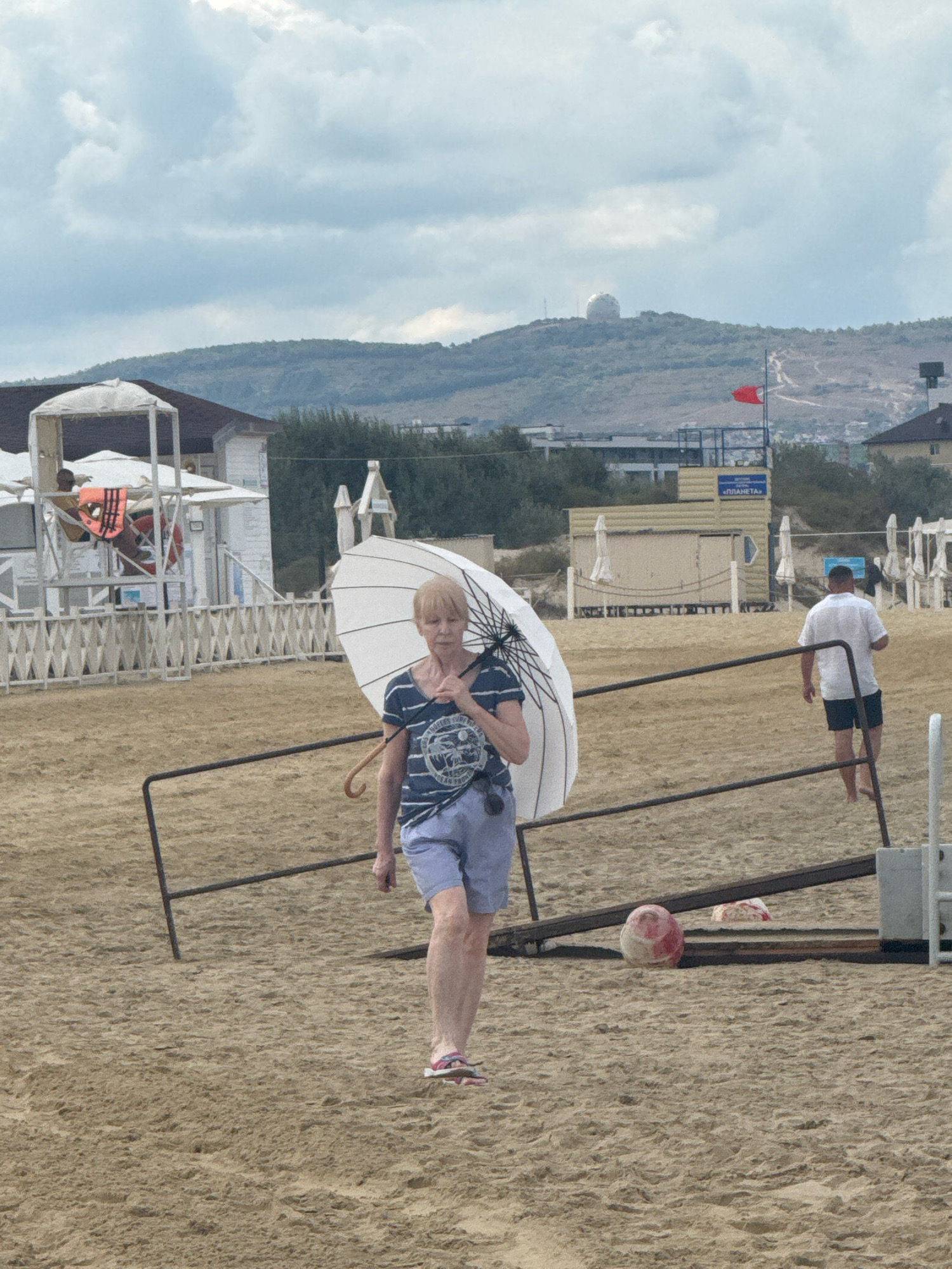 An elderly woman walks on a sandy beach in Anapa, Russia, holding a white umbrella. She wears a striped t-shirt and blue shorts. A man in a white shirt and black shorts walks away from her in the background. Several beach umbrellas and chairs are visible further down the beach, along with some structures. In the distance, a large spherical dome is visible on top of a hill. The sky is overcast.